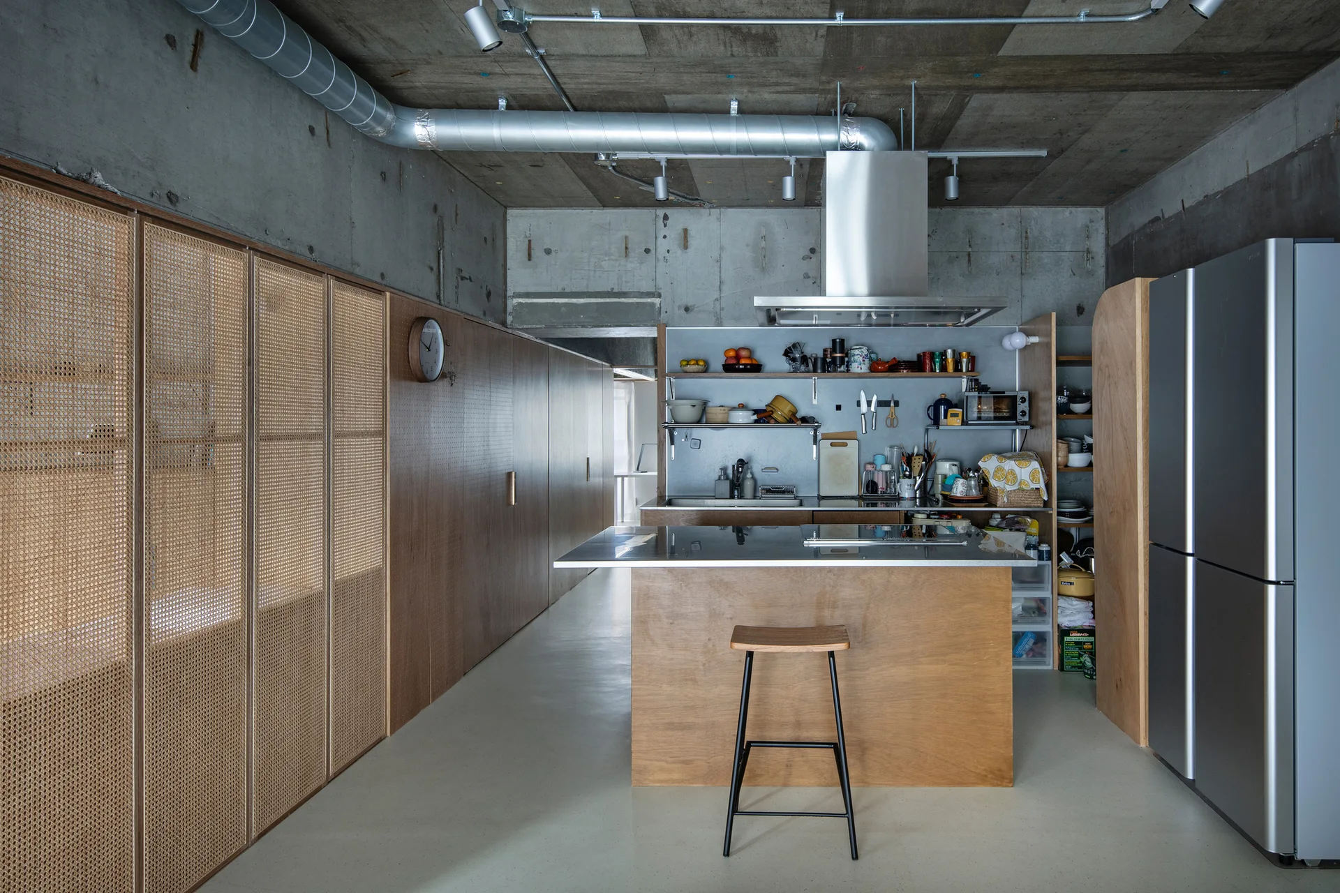 A modern industrial kitchen featuring an exposed concrete ceiling, a stainless steel kitchen island, and a long wall of wood and cane mesh cabinets.