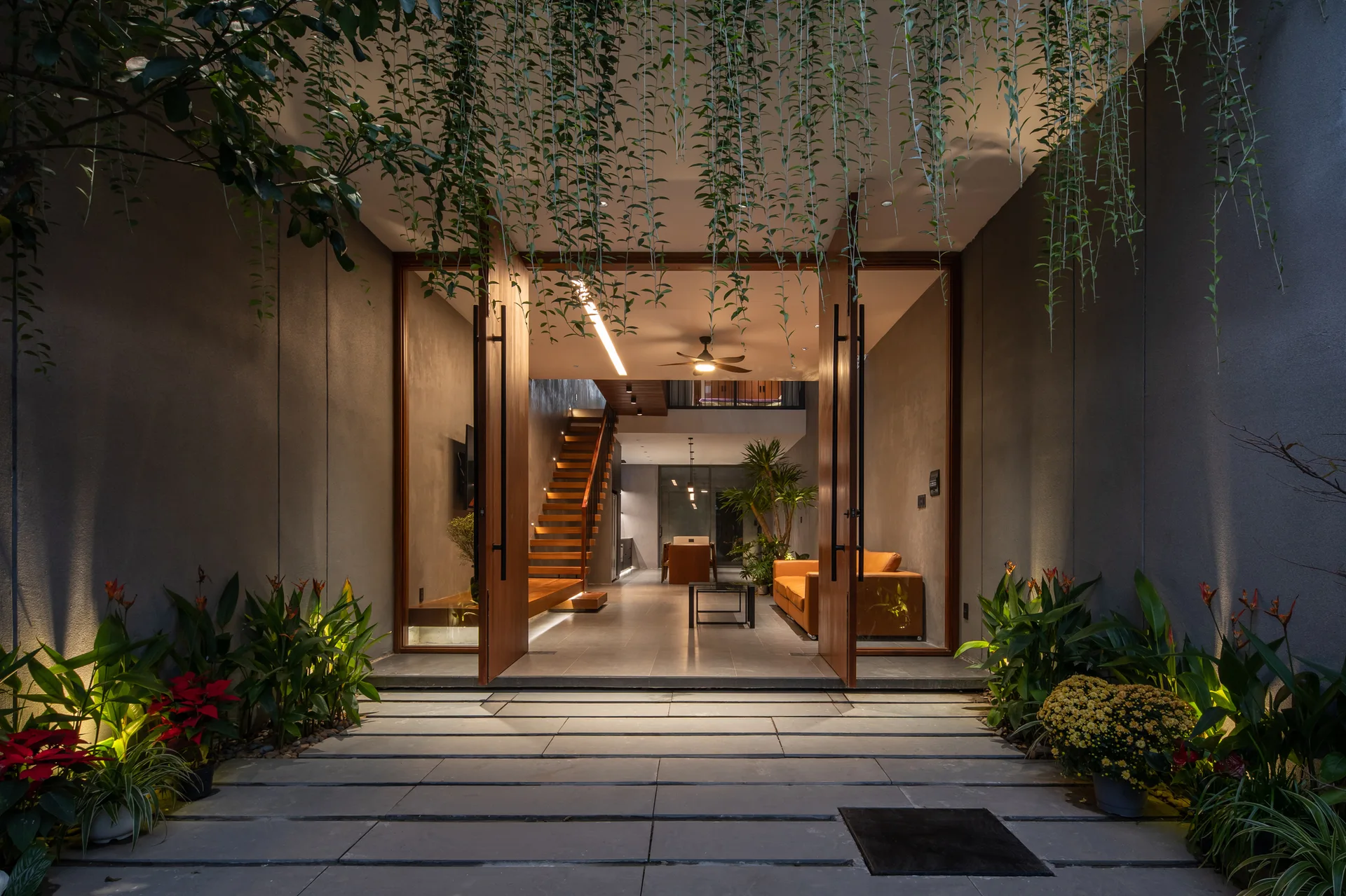 A stone pathway lined with plants leads through tall open wooden doors into a modern home with a wooden staircase and an orange sofa.