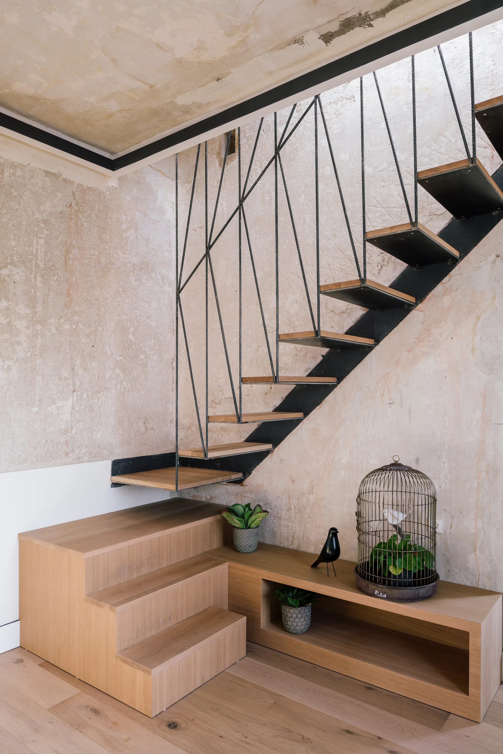 A floating staircase with wooden treads and a geometric black metal rod railing stands against a textured beige plaster wall above a light wood bench.