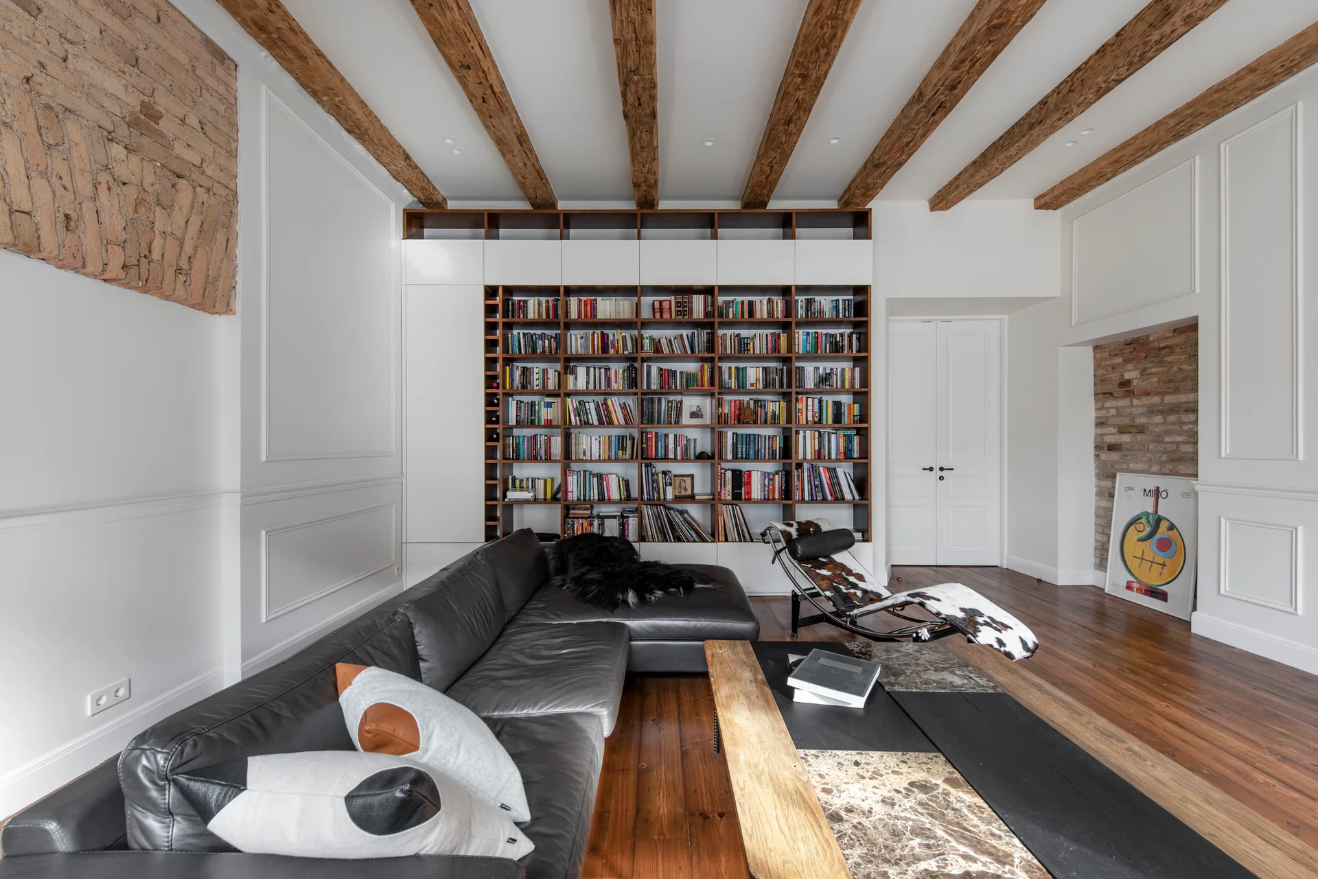 A living room features exposed timber ceiling beams, white paneled walls, a black leather sectional sofa, and a large built-in bookshelf.