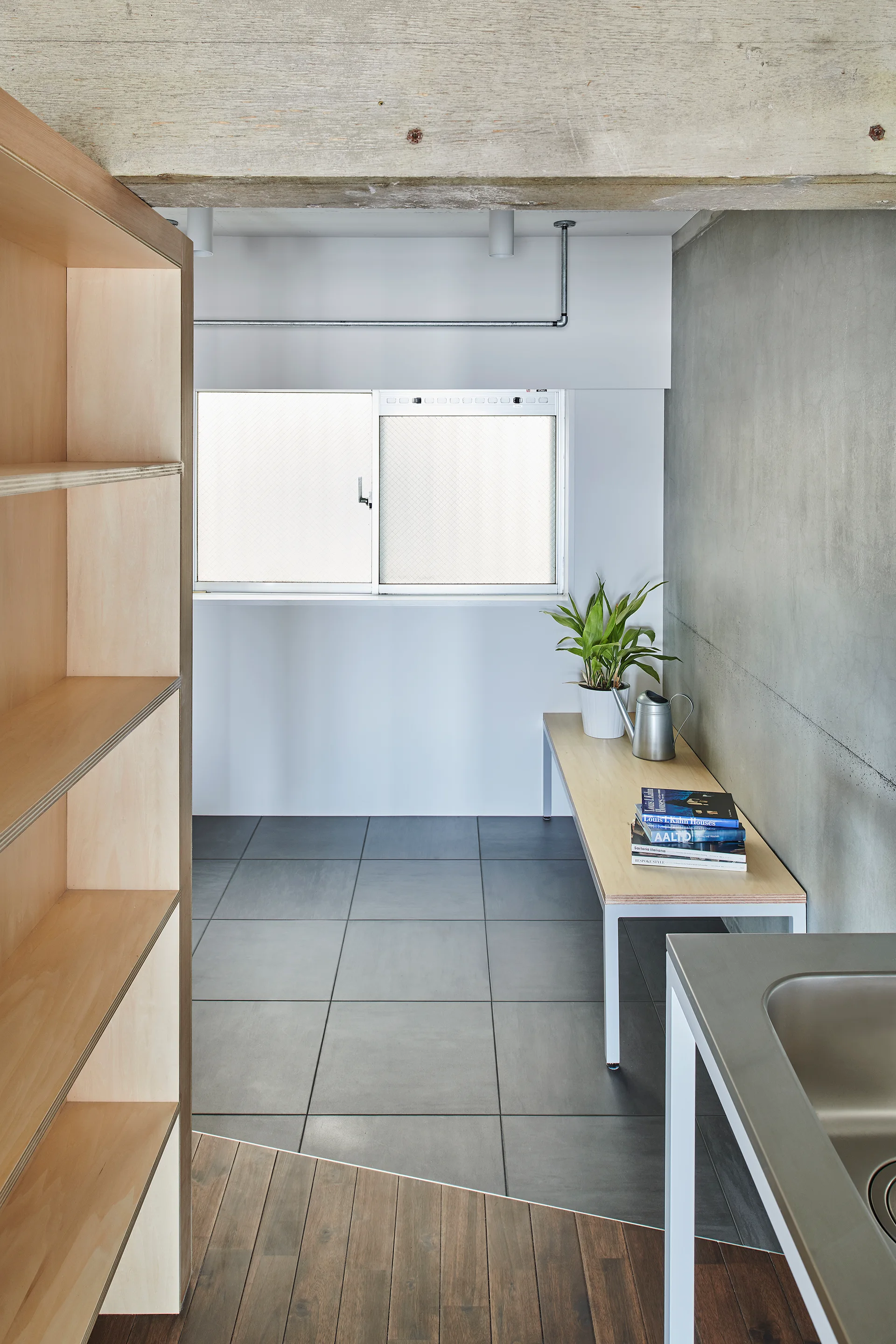A minimalist interior featuring an exposed concrete wall, a plywood shelving unit, grey floor tiles, and a small window above a wooden bench.