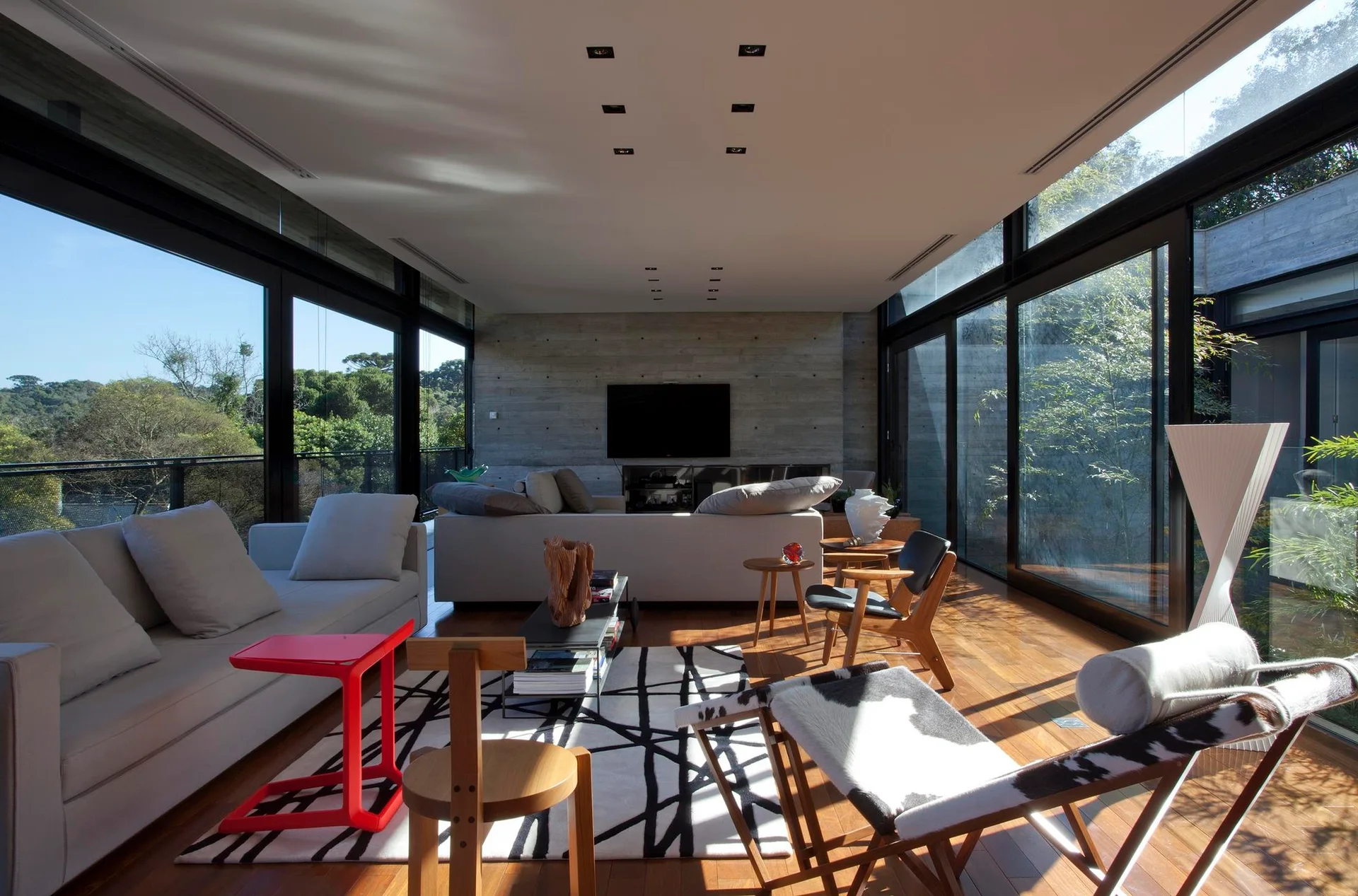 A modern living room featuring floor-to-ceiling glass walls, a textured concrete wall, wood flooring, grey sofas, and a graphic patterned rug.