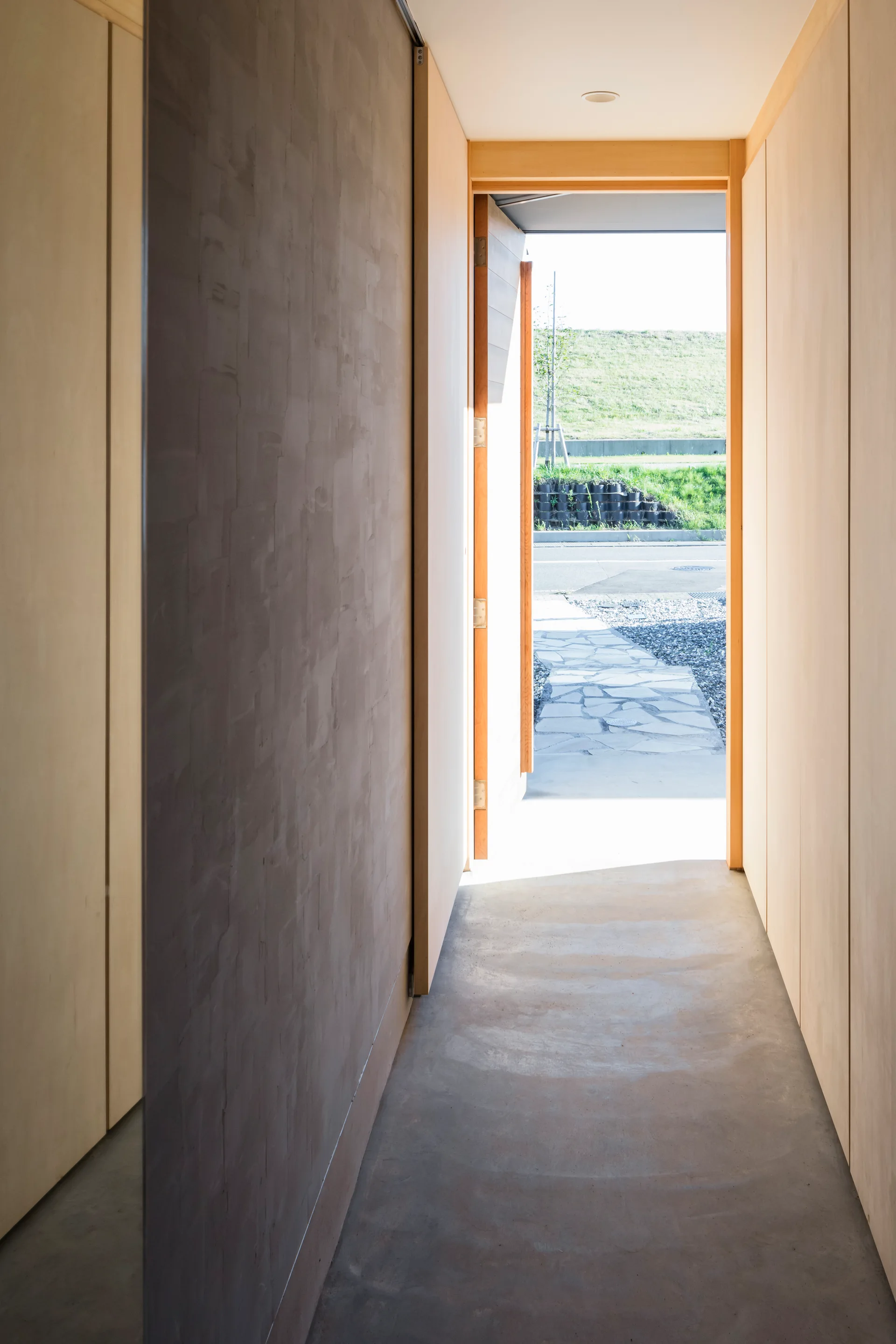 A narrow minimalist hallway features light wood paneled walls, a smooth concrete floor, and an open doorway looking out onto a stone path.