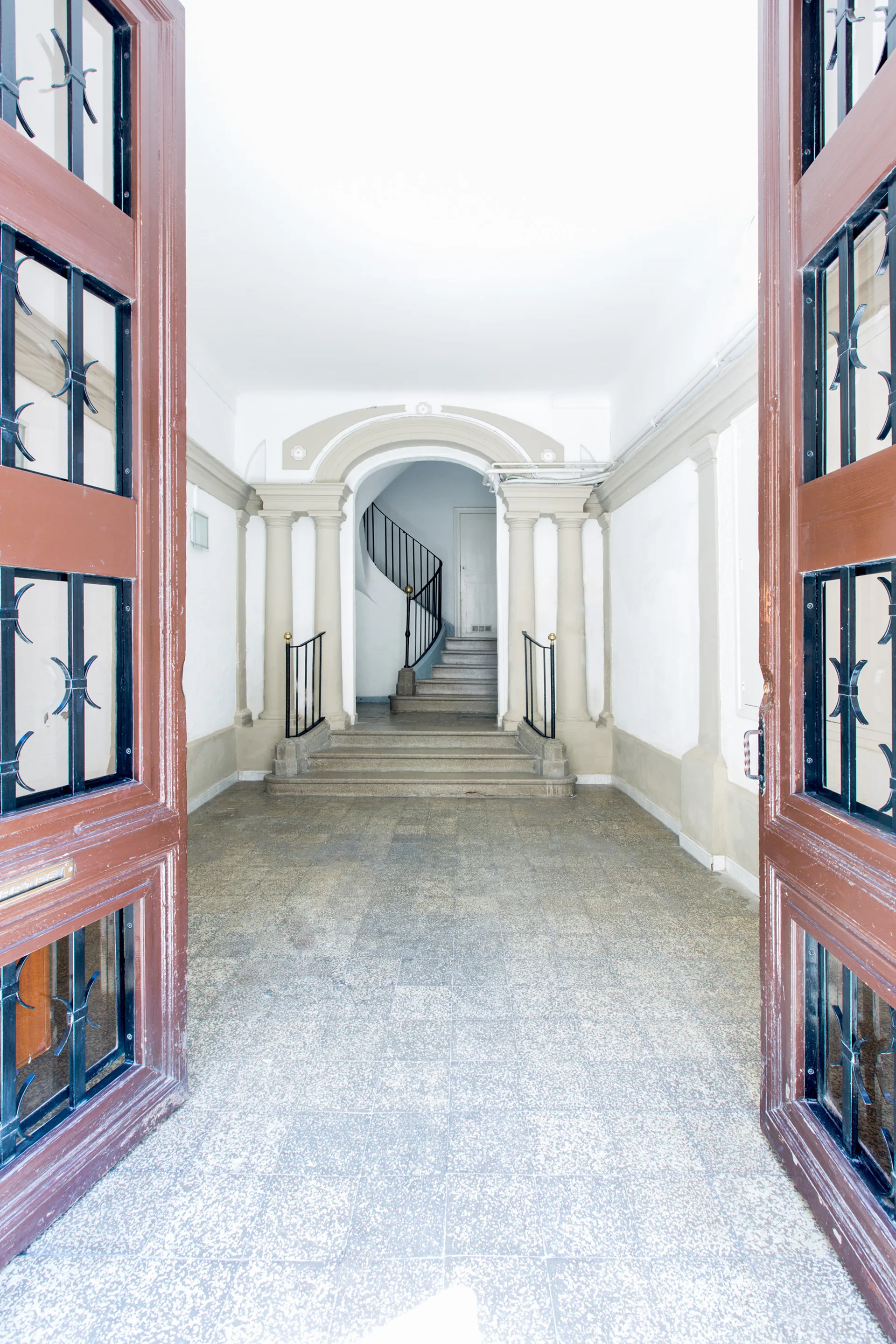Open wooden double doors with metal grilles look into a white neoclassical foyer with stone columns, an archway, and a curved staircase.
