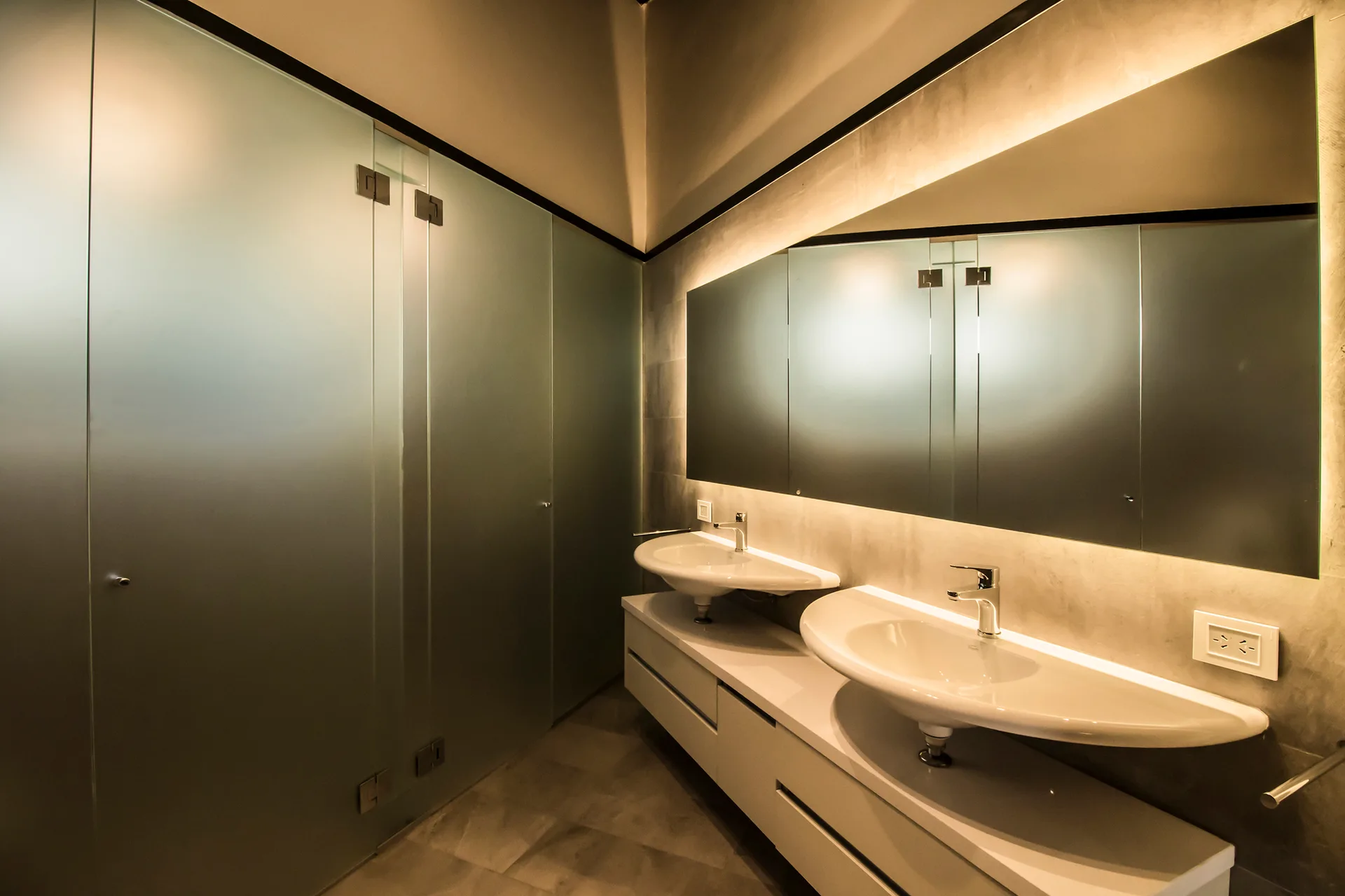 A contemporary gray bathroom features a white floating double vanity with two curved basins, a large backlit mirror, and frosted glass walls.