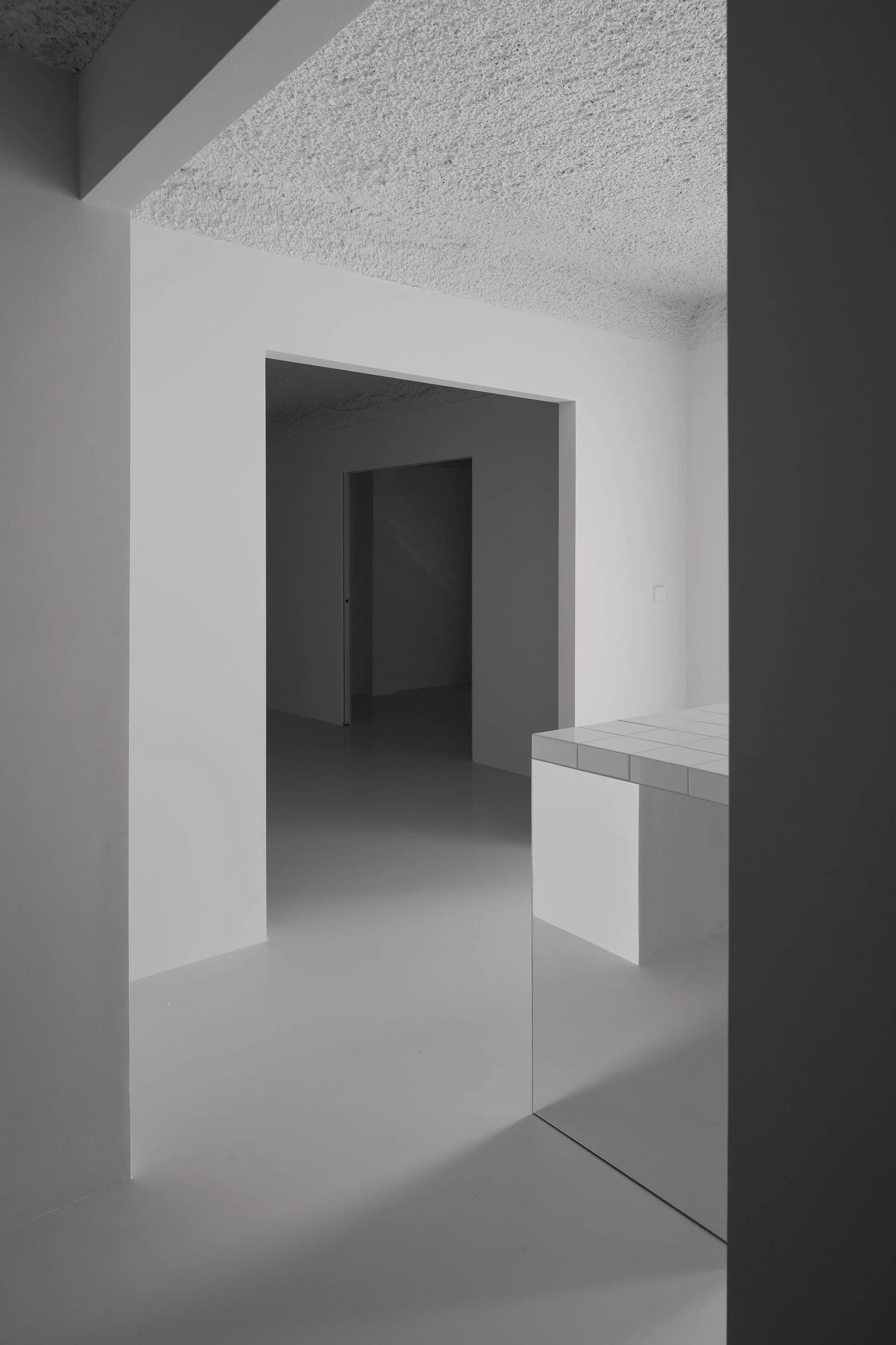 A monochromatic white minimalist kitchen featuring a tiled counter island, seamless flooring, a heavily textured ceiling, and nested square doorways.