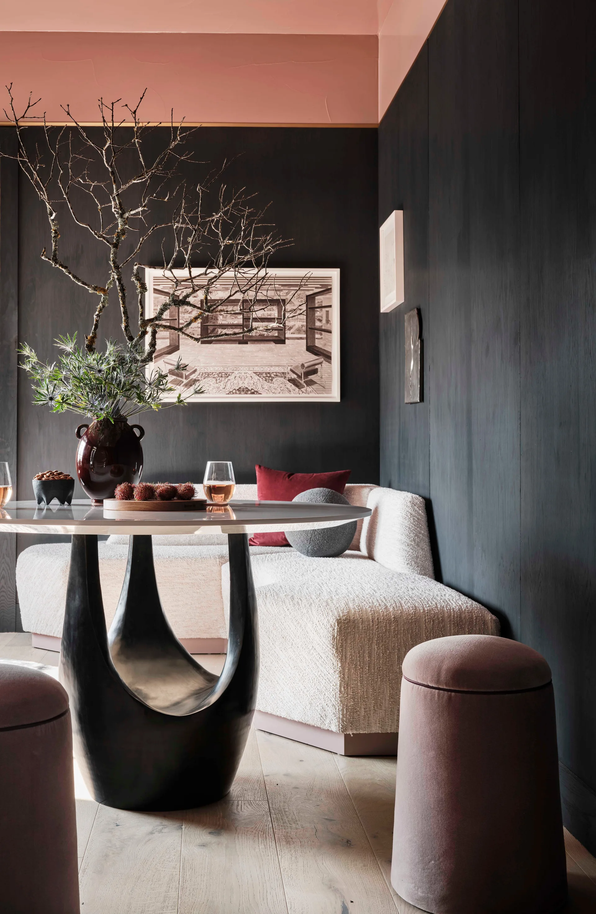 A modern dining nook features dark charcoal wood paneled walls, a textured cream banquette, and a circular marble table beneath a blush pink ceiling.