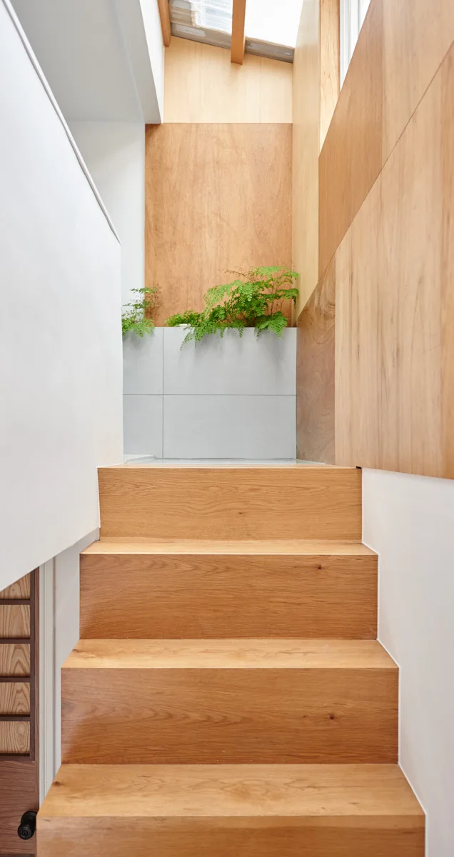 A wooden staircase leads to a landing with a grey built-in planter and wood-paneled walls illuminated by an overhead skylight.