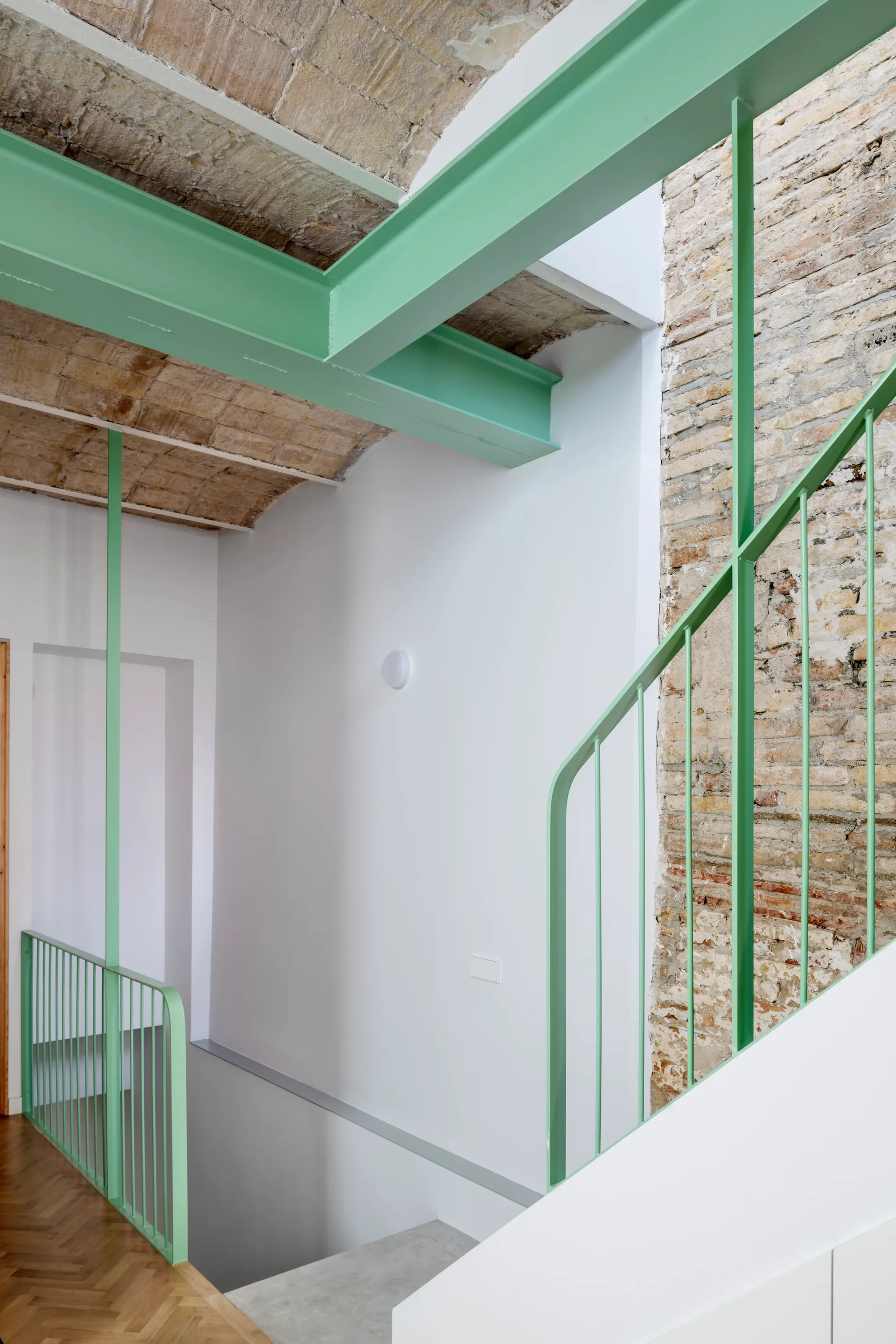 A mint green metal staircase railing and steel ceiling beams stand against white walls and an exposed brick vaulted ceiling.