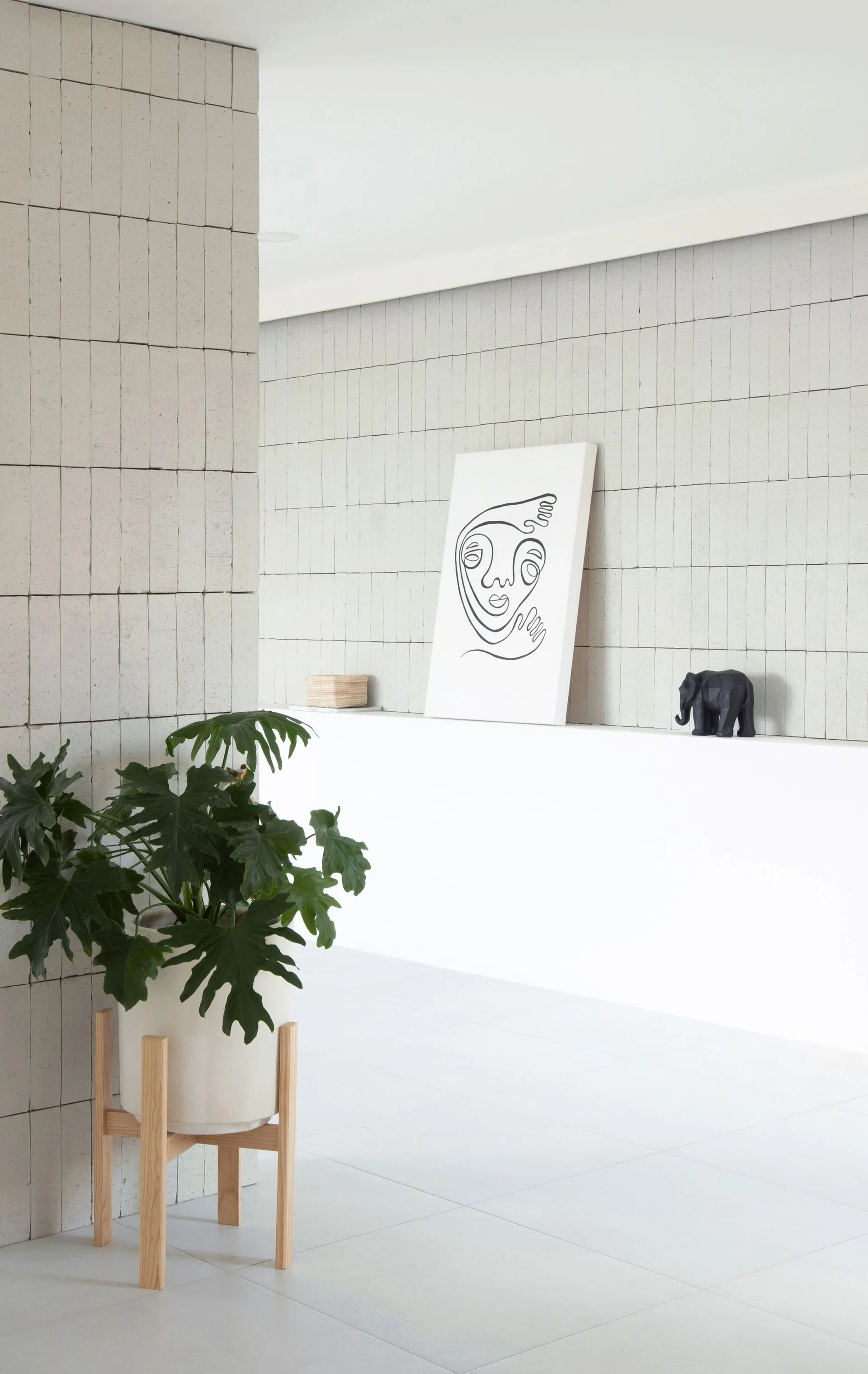 A bright minimalist interior featuring a light gray tiled masonry wall, a white ledge displaying line art, and a potted plant on a wooden stand.