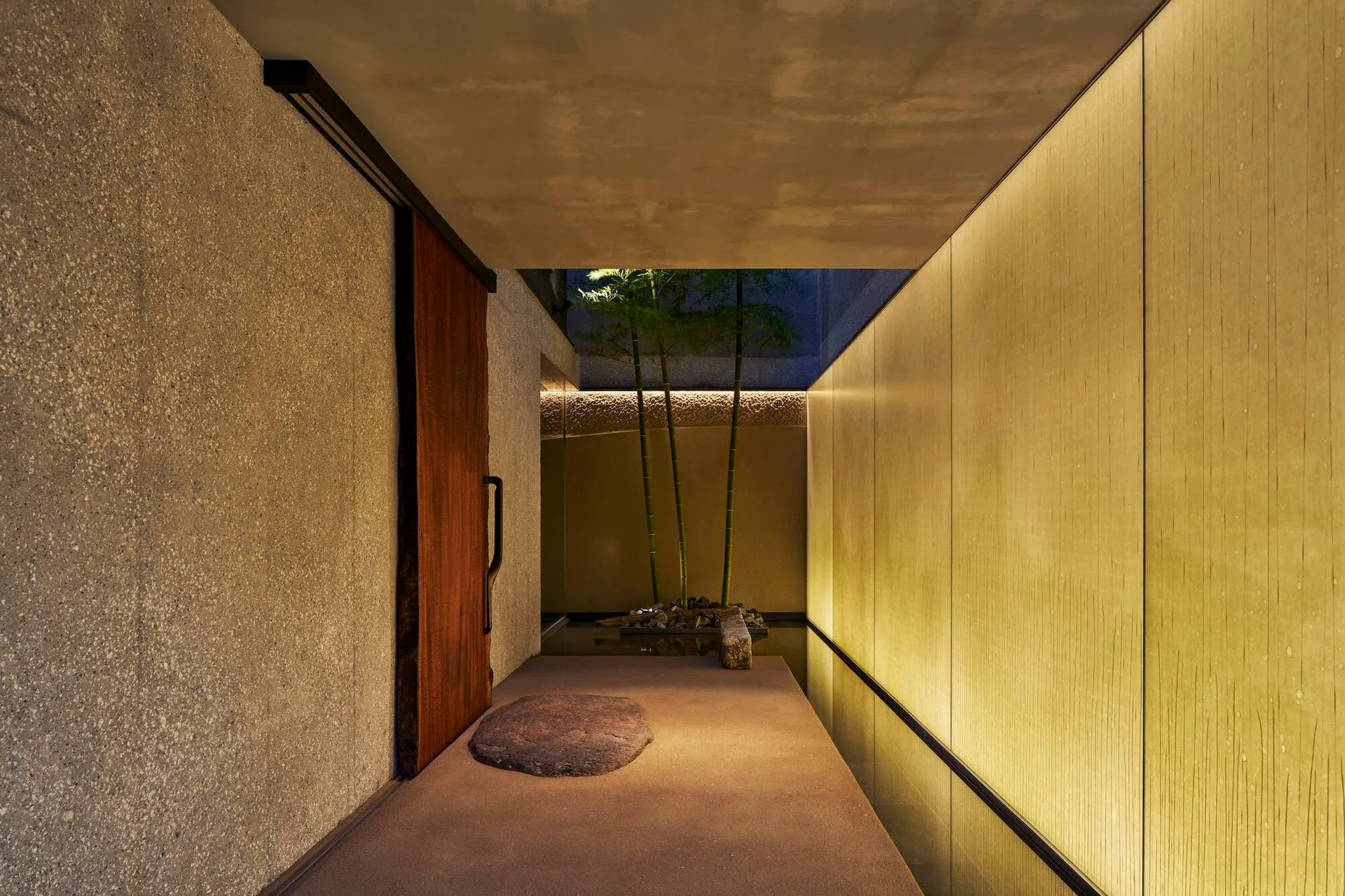 A minimalist entryway featuring a rustic sliding wood door, textured stone walls, and a glowing gold wall leading to a bamboo courtyard.