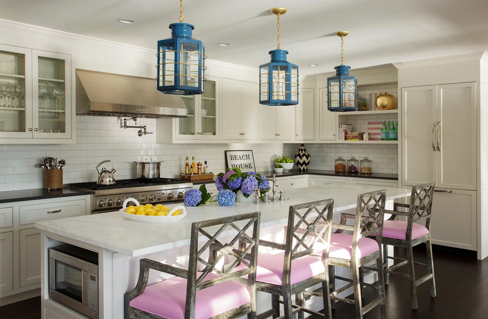 A white kitchen features a marble-topped island with pink-cushioned wooden stools beneath three hanging blue lantern pendant lights.