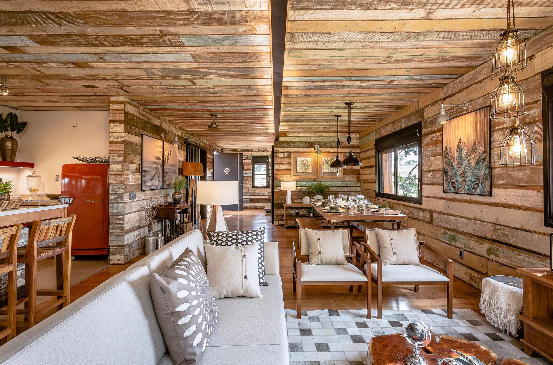 An open-concept living space features walls and a ceiling clad in weathered reclaimed wood, furnished with a light gray sofa and wooden dining set.