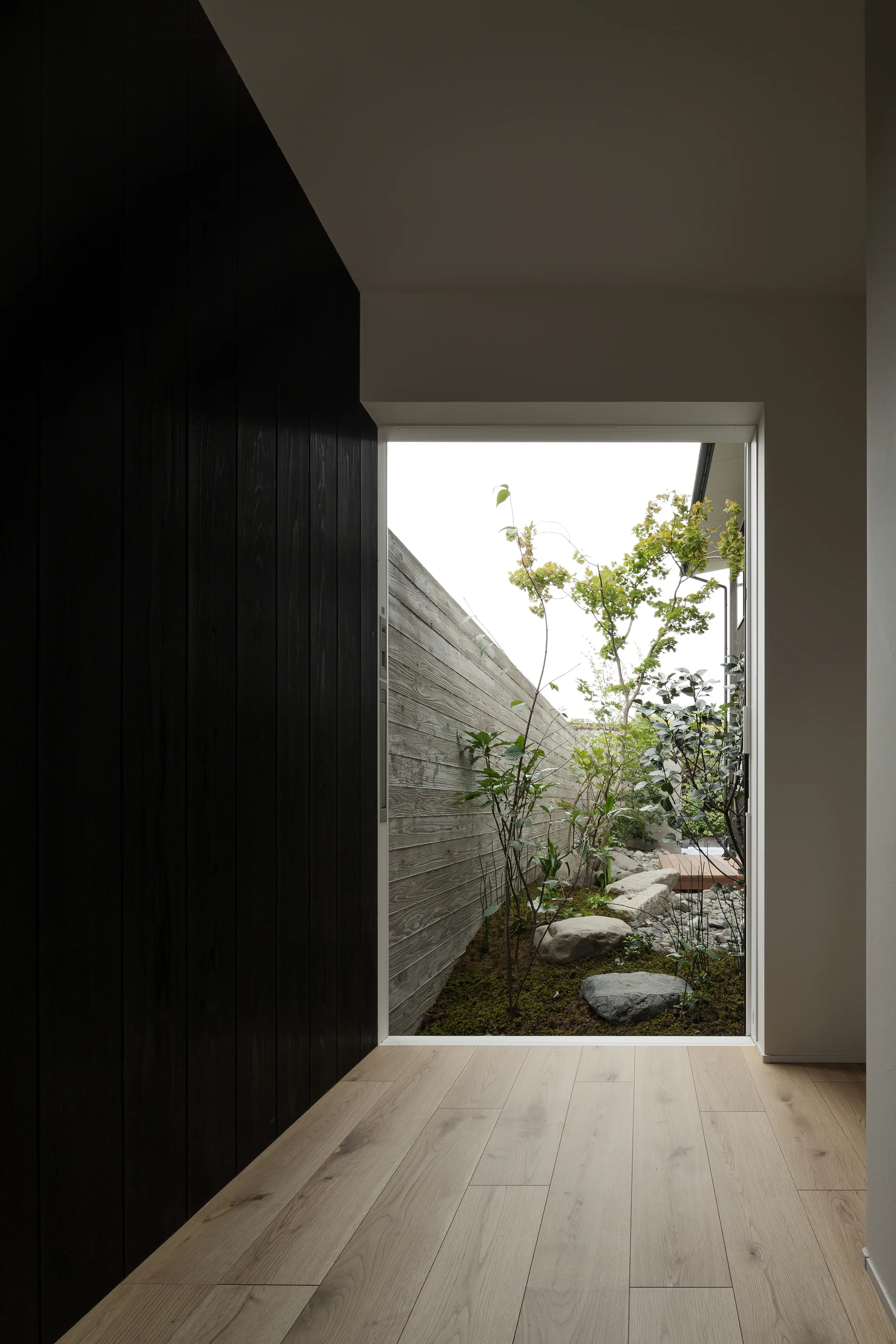 A minimalist hallway features light wood floors and a dark paneled wall framing a view of an outdoor courtyard with stones and trees.