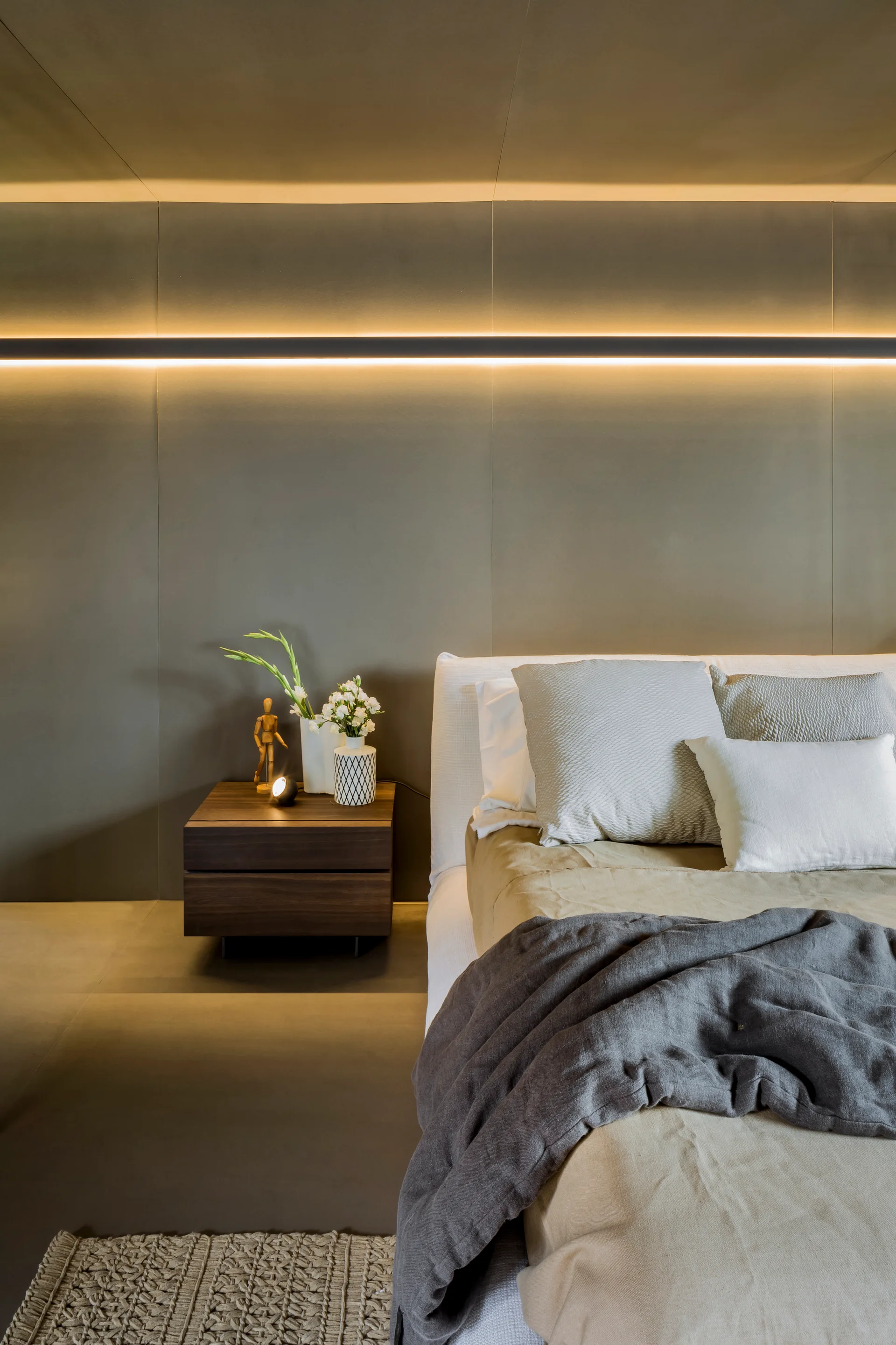 A neutral bedroom features a bed with layered linens next to a floating wooden nightstand against a gray paneled wall with an integrated warm light strip.