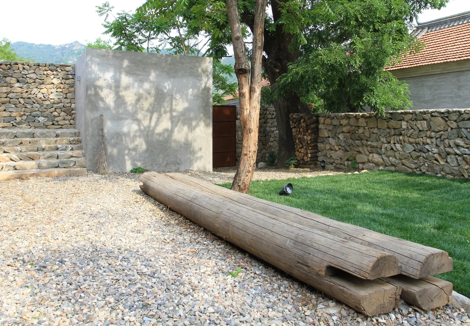 A rustic courtyard featuring a dry-stacked stone wall, a smooth concrete structure, steps, and a large raw timber log bench on gravel.