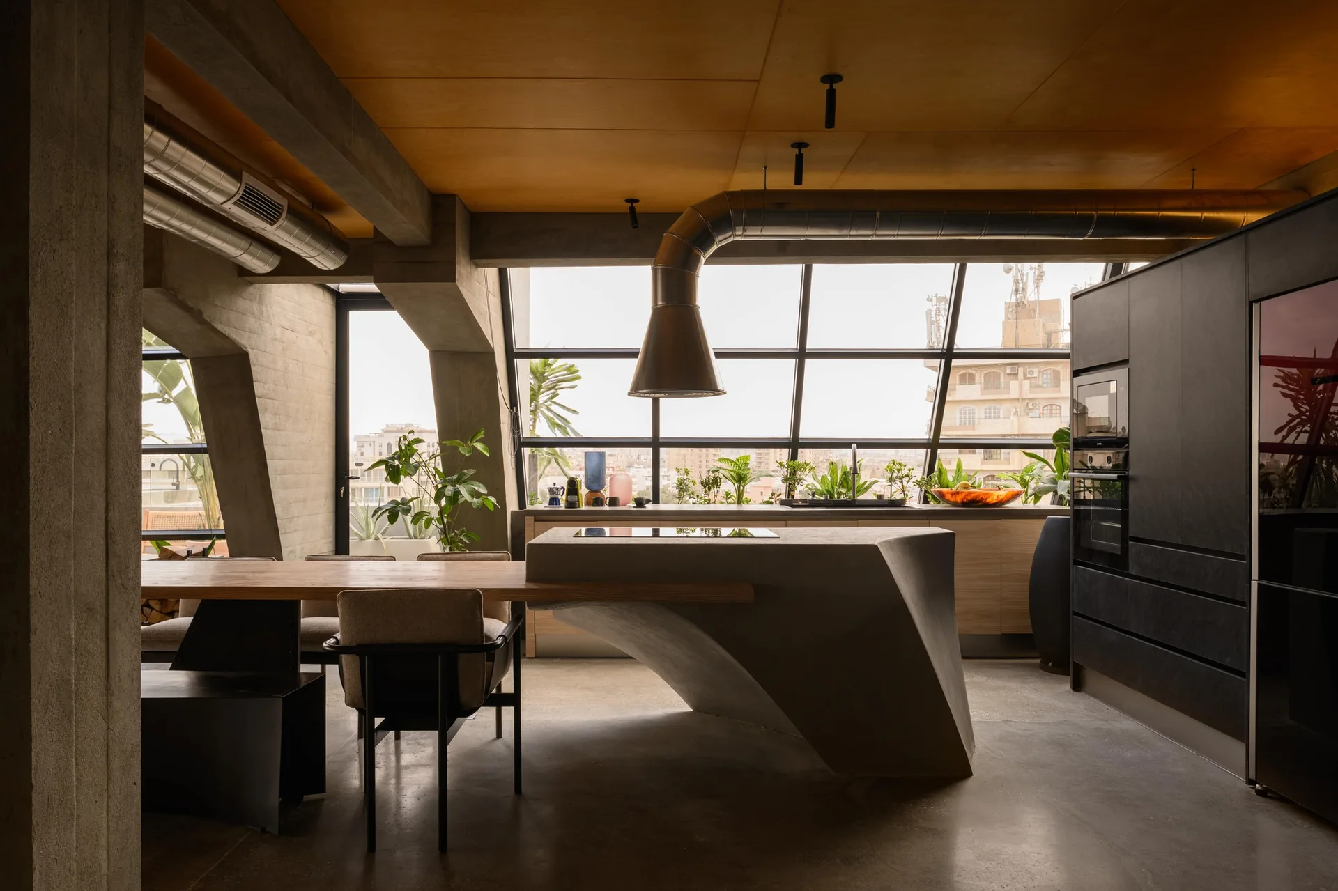 An industrial kitchen showcases a sculptural concrete island connected to a wooden dining table near a large slanted window wall and concrete beams.