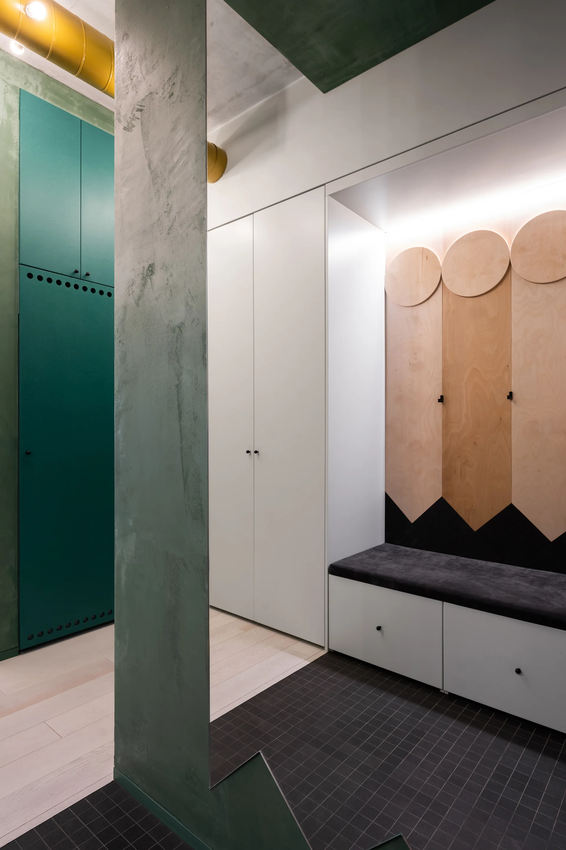 A modern mudroom entryway features built-in white cabinets, a cushioned storage bench with geometric wood paneling, and a textured green column.