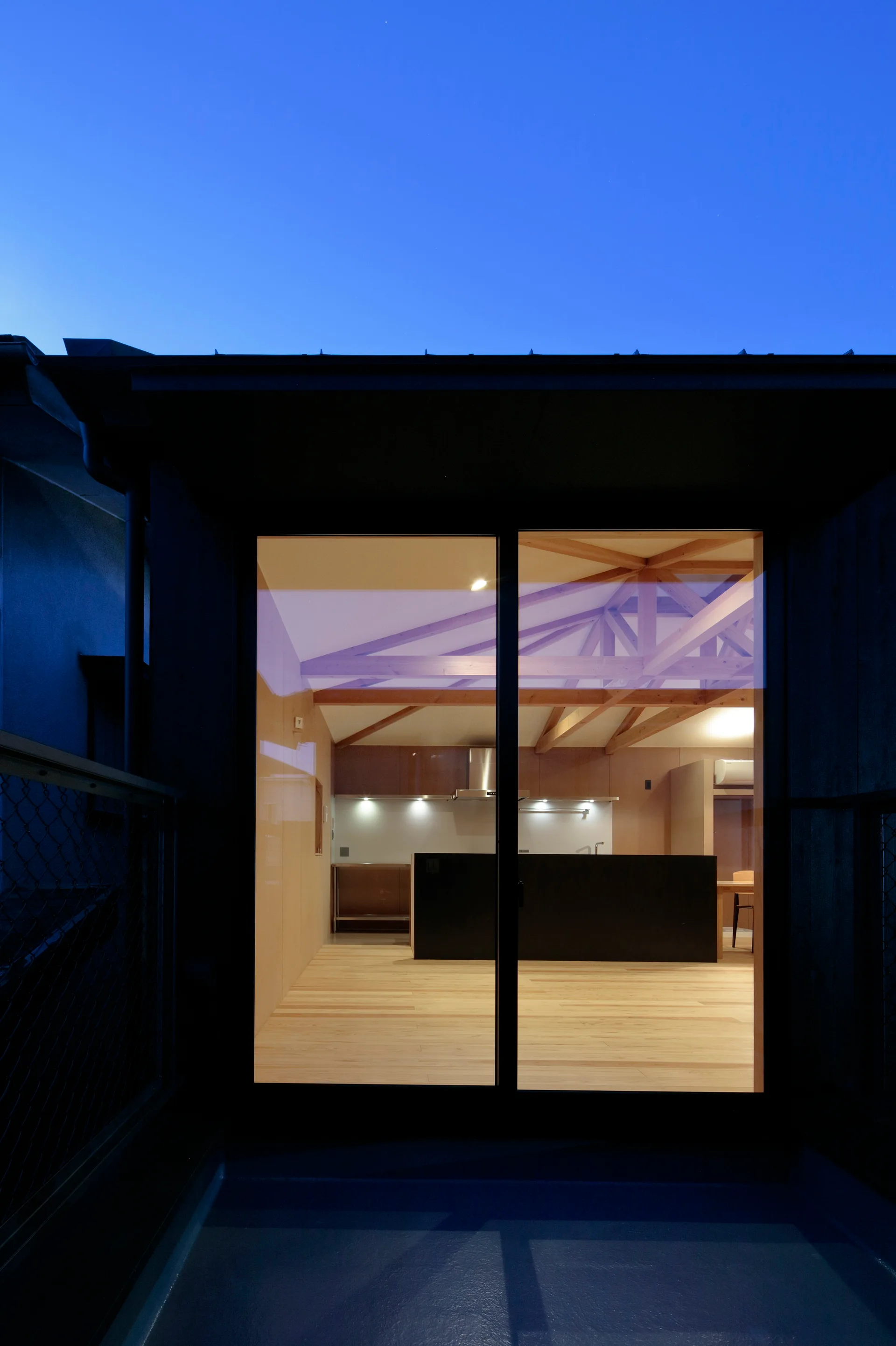A twilight view through a sliding glass door reveals a minimalist kitchen with a black island, light wood floors, and exposed wooden roof trusses.