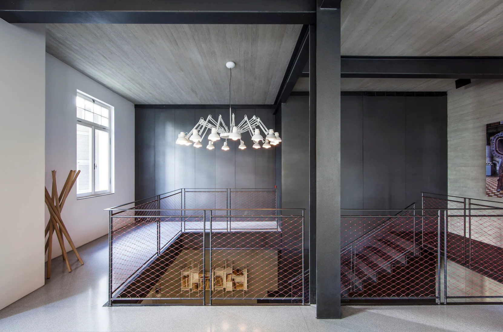 An industrial stairwell landing features a black metal railing filled with red mesh netting, a large white multi-arm chandelier, and exposed steel columns.