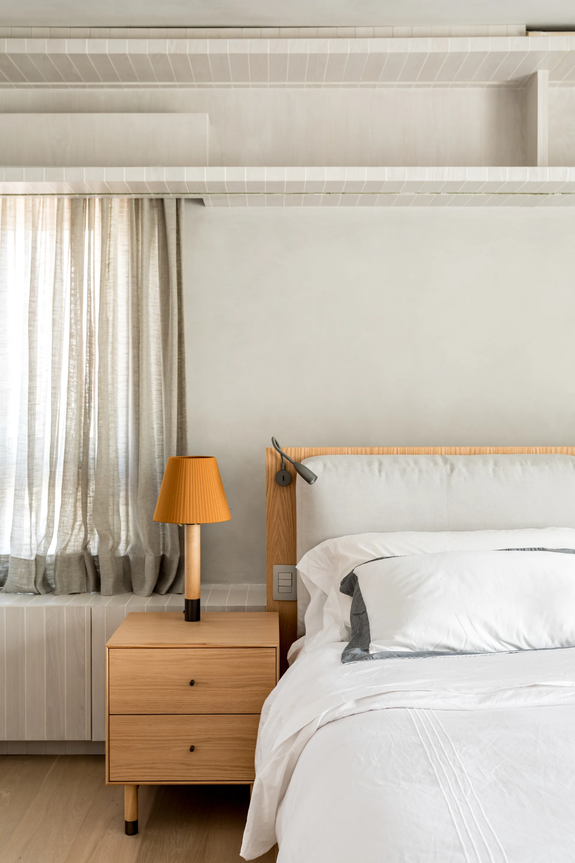 A minimalist bedroom featuring a wooden headboard, a light oak bedside drawer with an orange pleated lamp, and built-in wall shelving.
