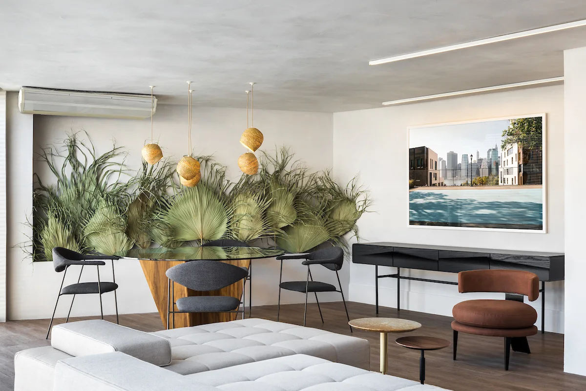 A modern open dining and living space featuring a glass-top table, dark chairs, a black sideboard, and a wall alcove filled with dried palm leaves.