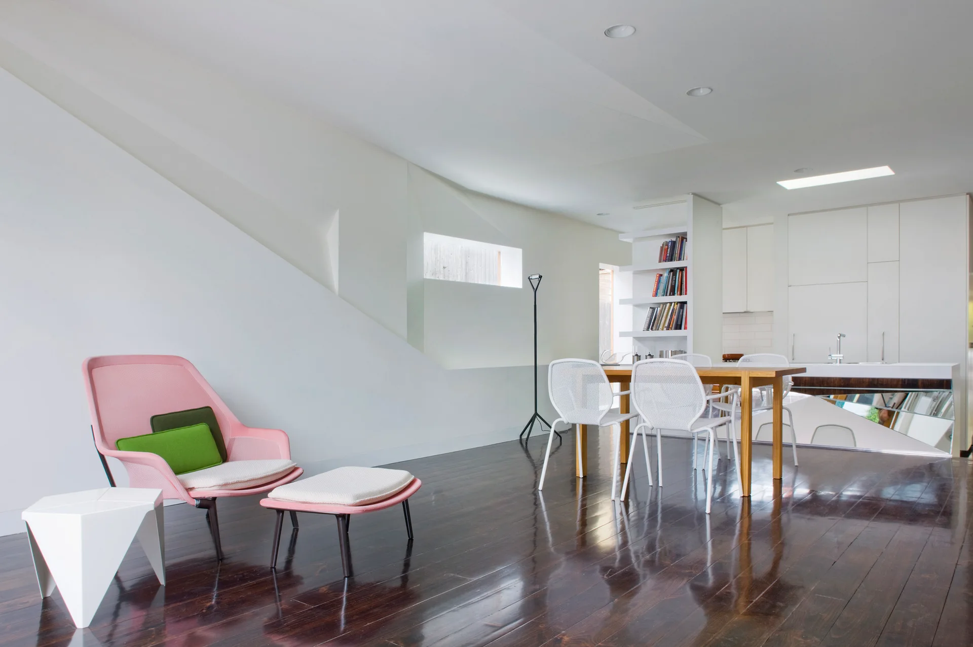 A pink lounge chair and ottoman sit on dark glossy hardwood floors next to a white geometric side table in an open-plan living room.