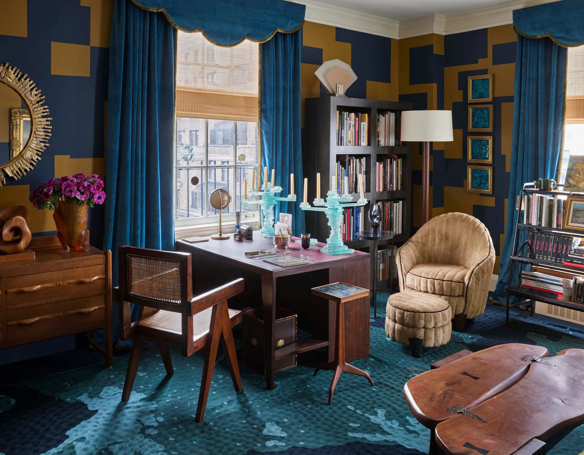 A maximalist home office features a dark wood desk with a cane chair, blue velvet curtains with a scalloped valance, and geometric blue and gold wallpaper.