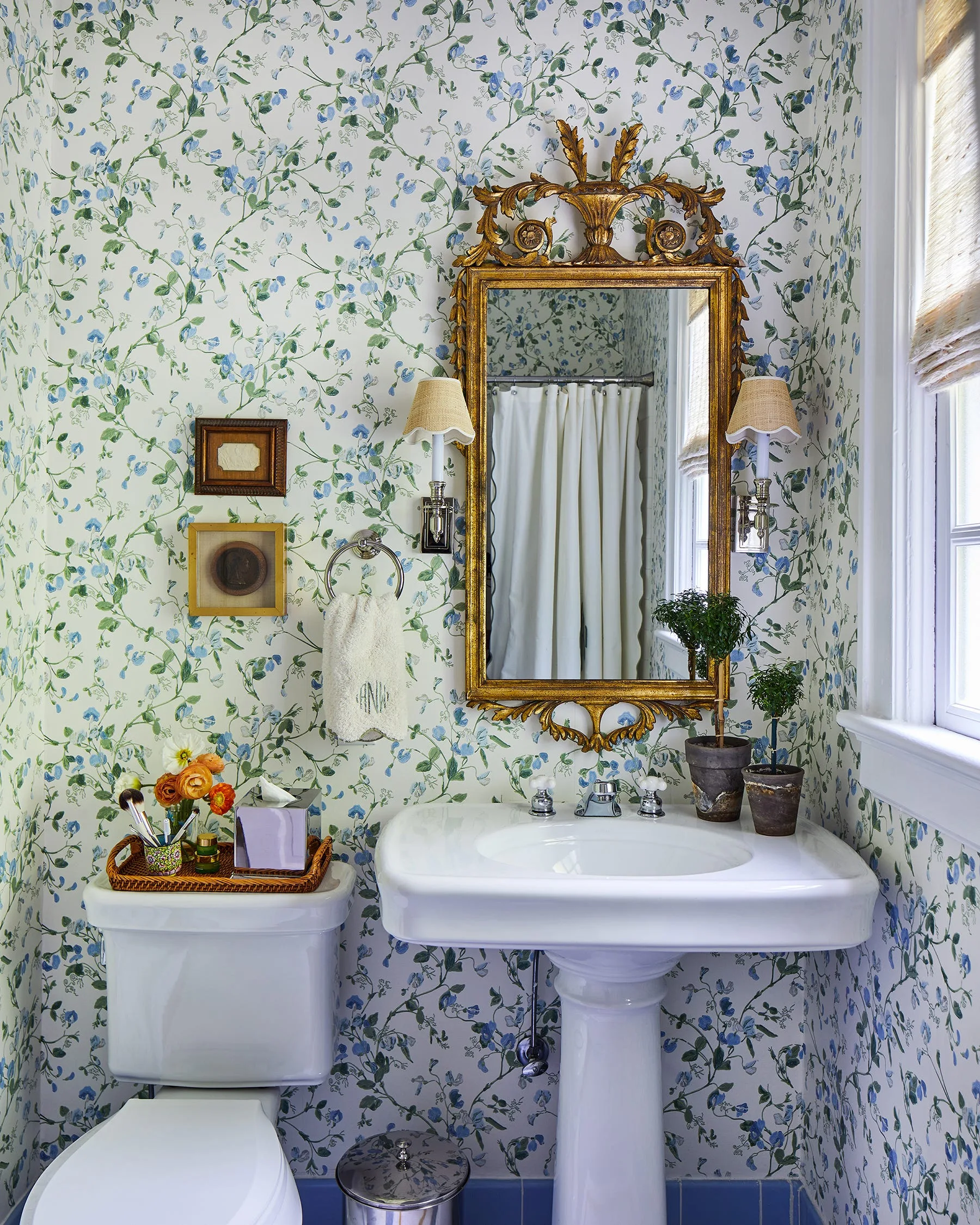A powder room with blue and white floral wallpaper, a white pedestal sink, and an ornate gold mirror flanked by two wall sconces.