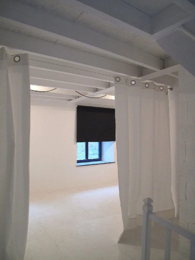 Minimalist white room divider curtains hang below painted ceiling joists opposite a window with a stark black roller blind.