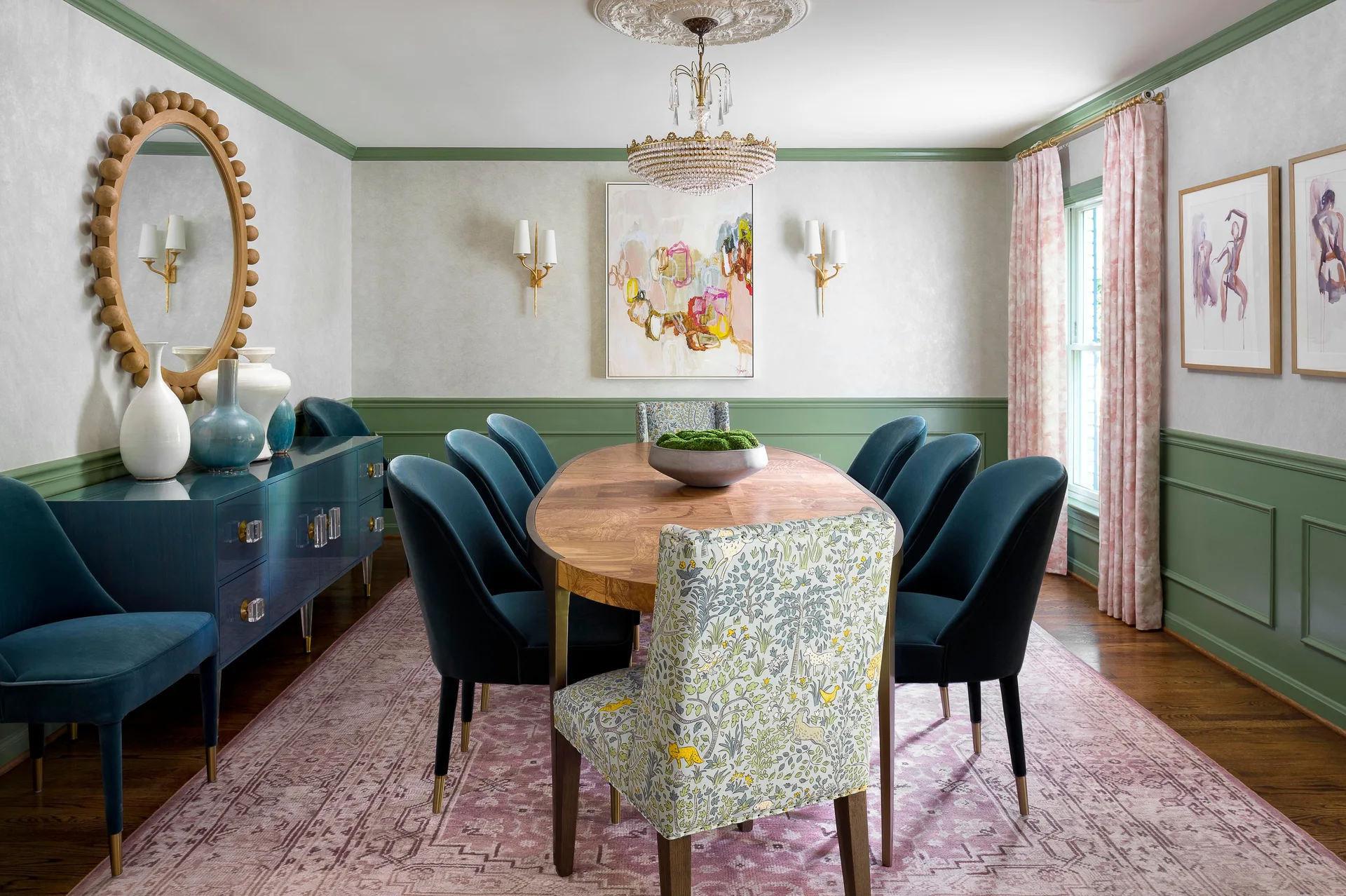 A formal dining room features an oval wood table, blue velvet chairs, green wainscoting, a crystal chandelier, and a patterned pink area rug.