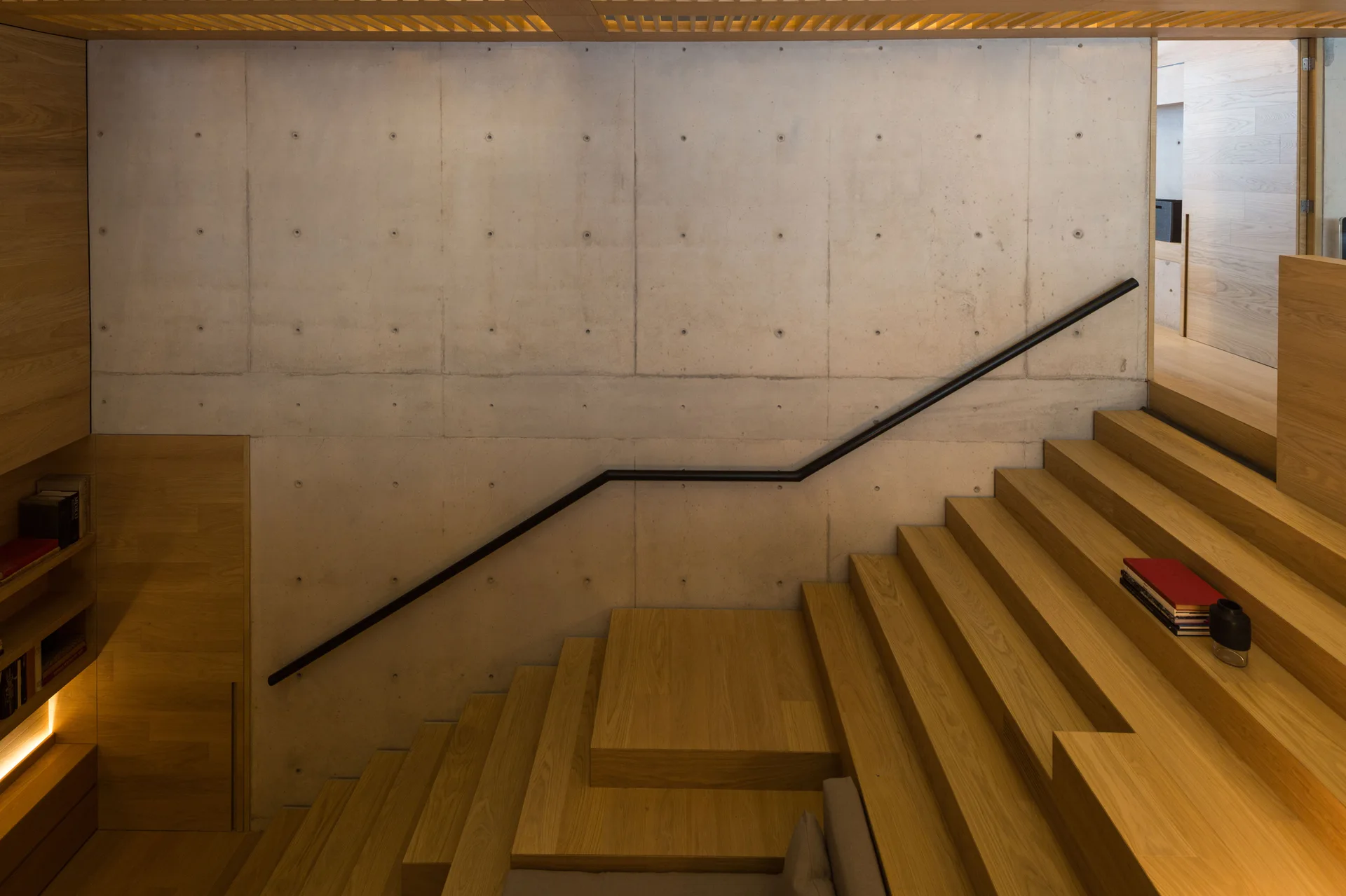 A wide wooden staircase with integrated shelving rises against a large gray concrete wall with a black metal handrail.