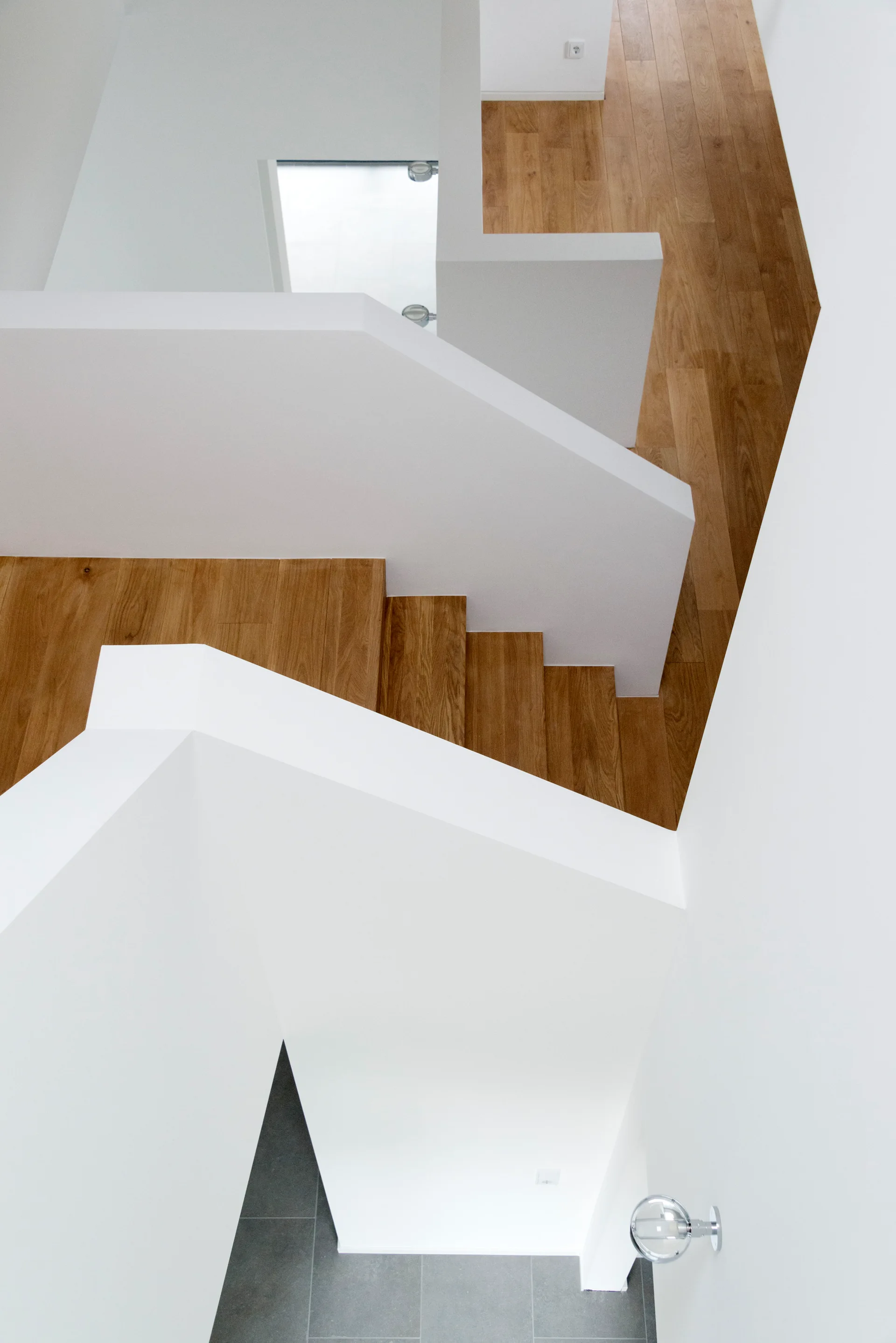 A high-angle overhead view of a minimalist multi-level staircase featuring medium-tone wooden steps and solid, thick white plaster railings.