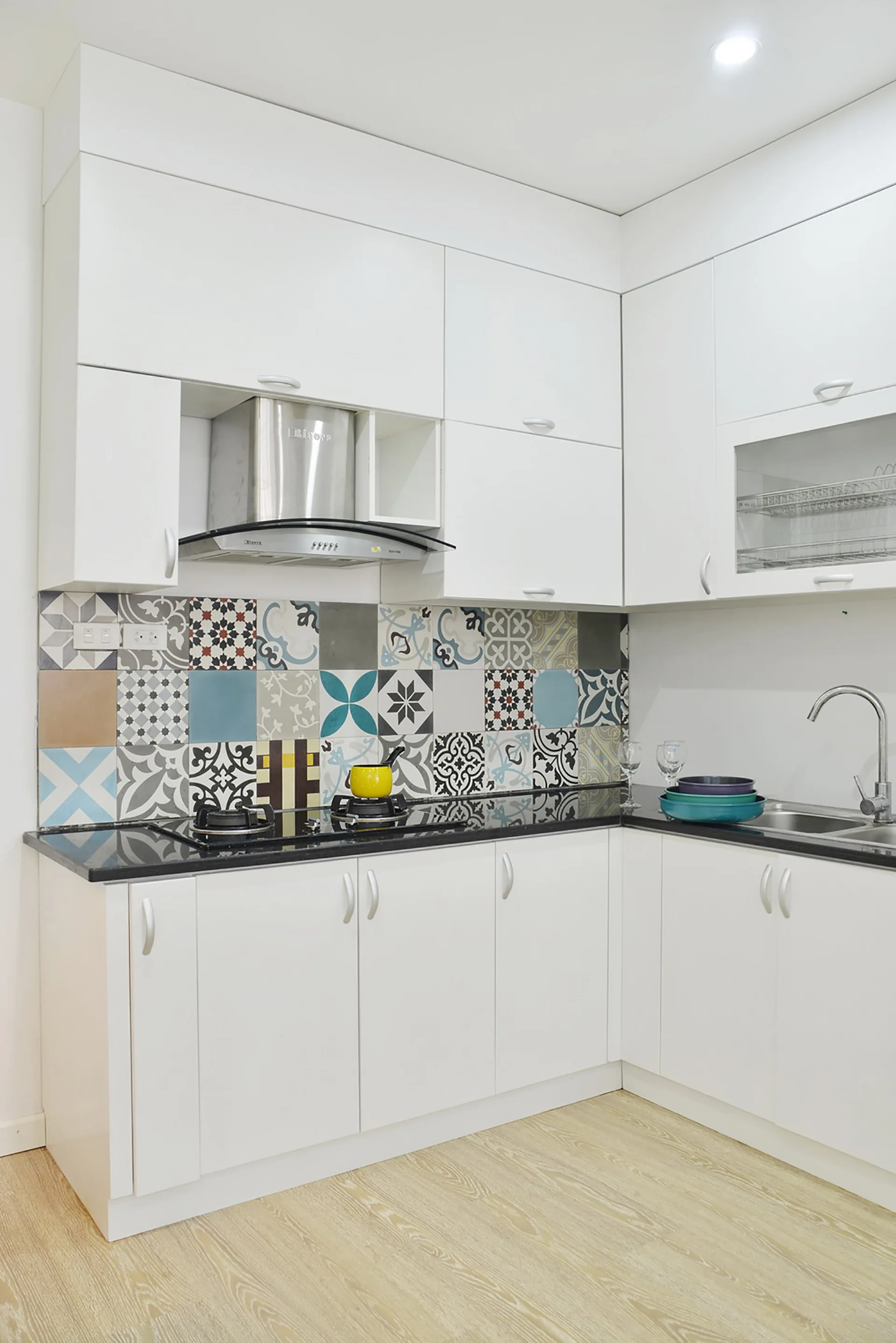 A modern kitchen corner features white flat-panel cabinetry, a black countertop, and a colorful patterned tile backsplash.