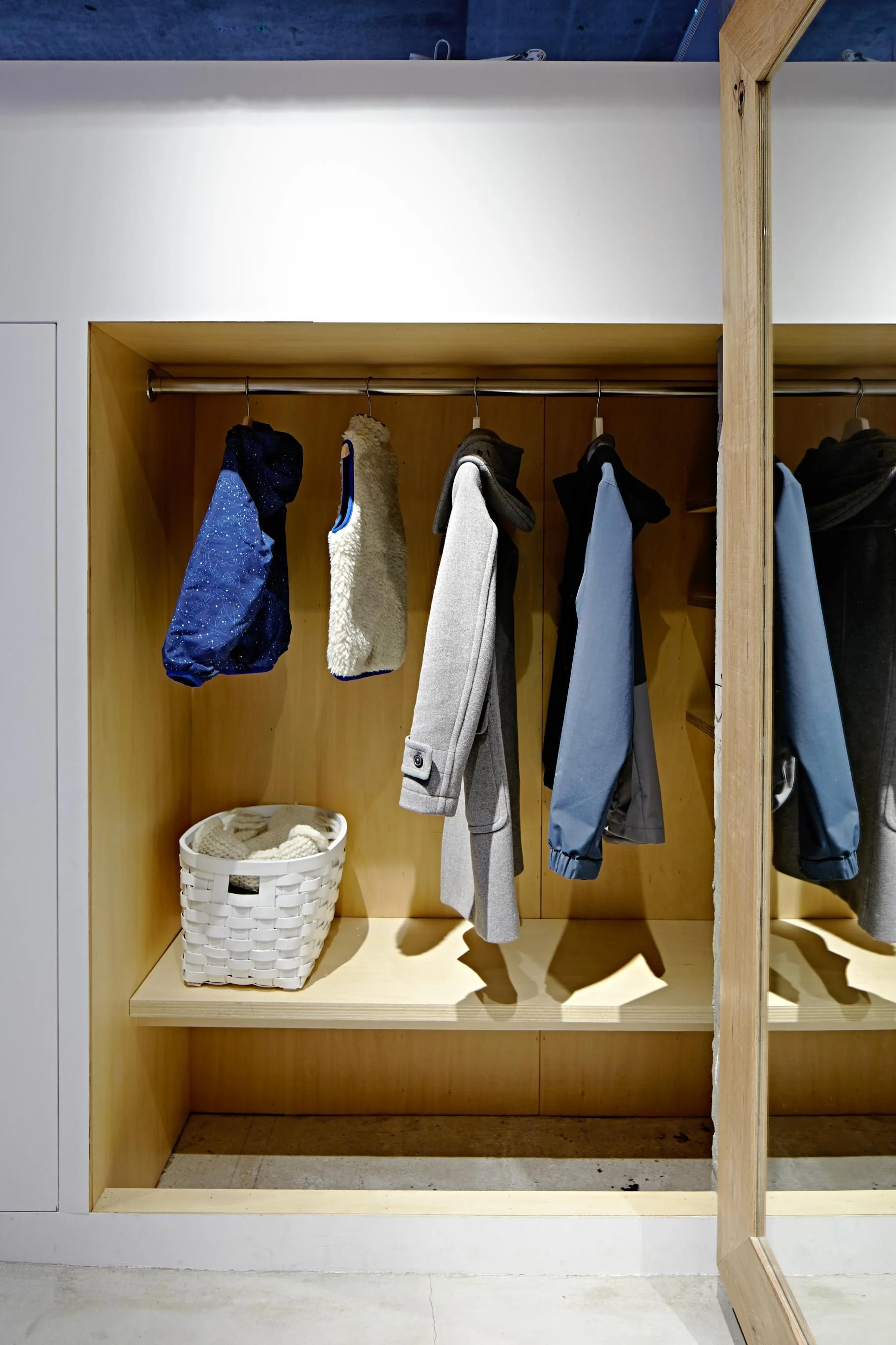 A recessed light wood closet features a hanging rod with winter coats, a lower shelf holding a white woven basket, and a matching wood-framed mirror.