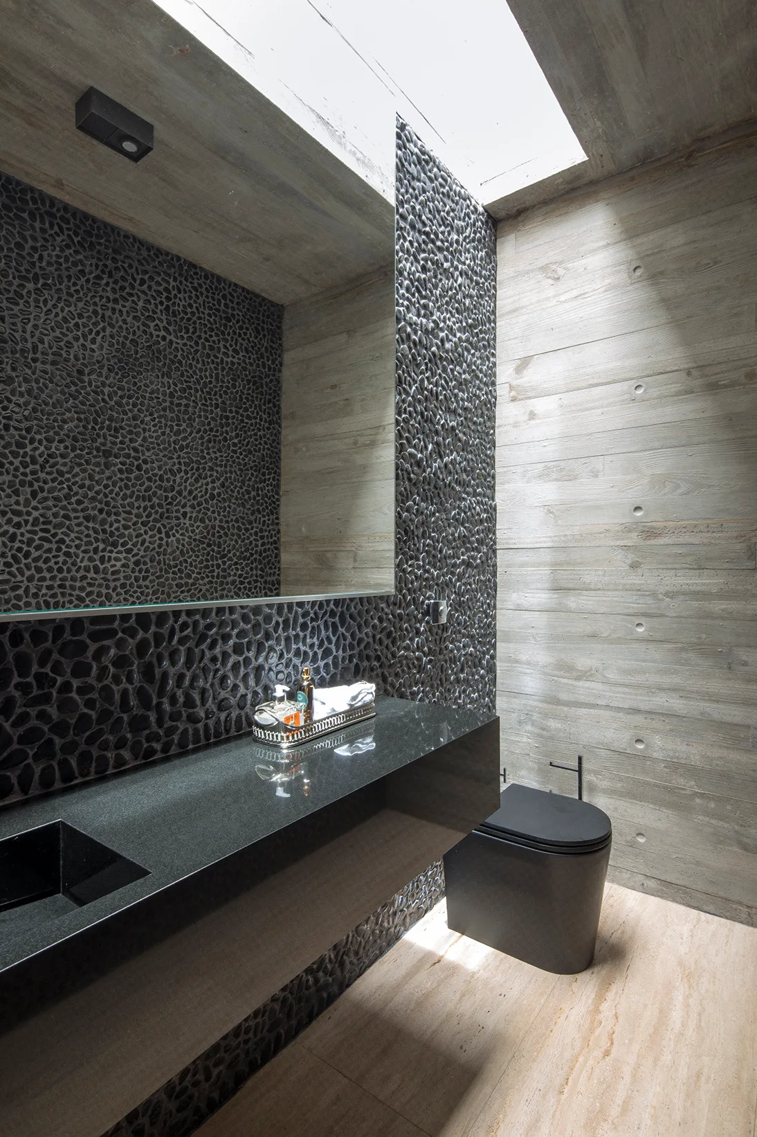 A minimalist powder room featuring a black floating vanity, textured pebble wall, matte black toilet, and raw concrete walls illuminated by a skylight.