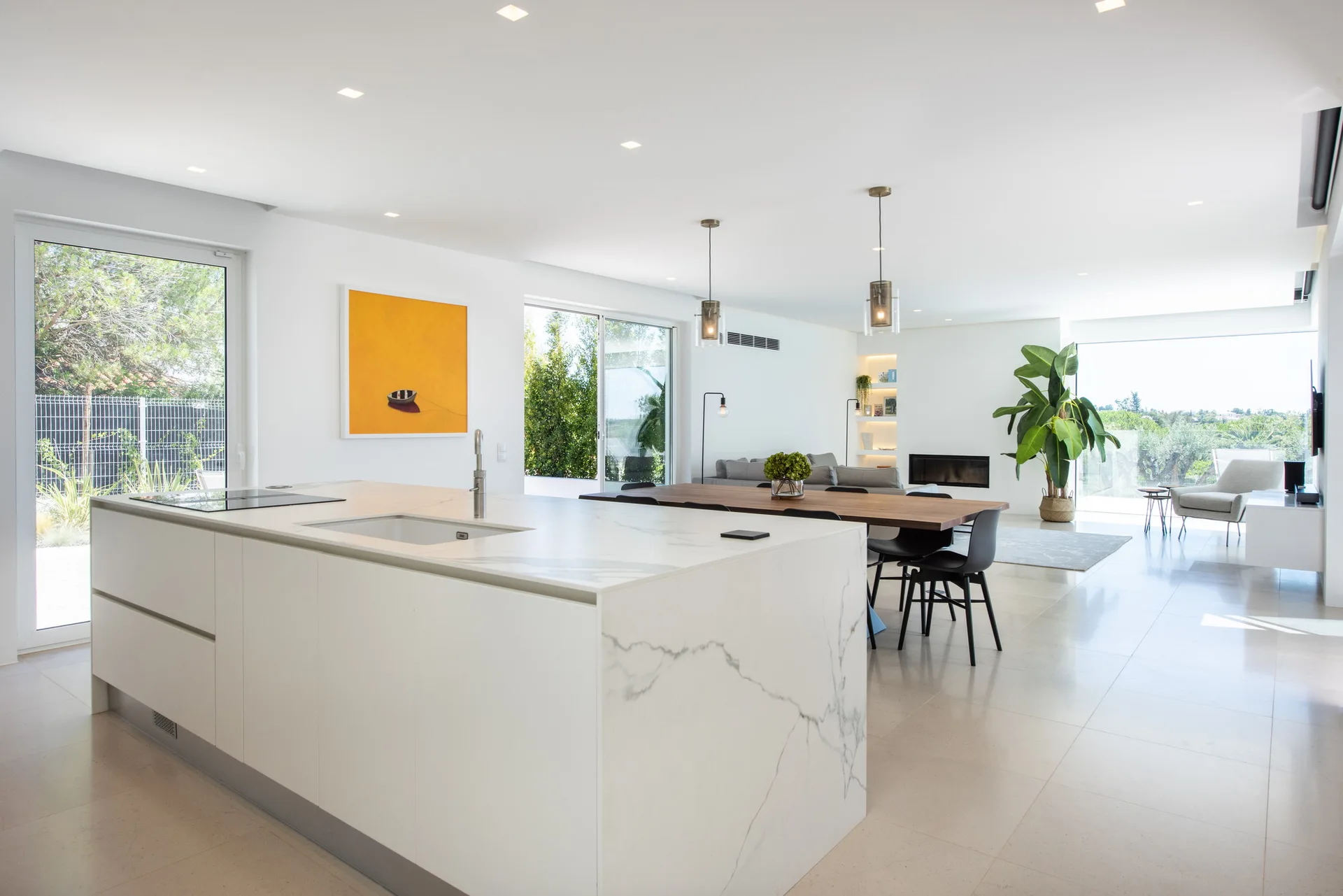 A modern open-plan kitchen features a white marble waterfall island, a wooden dining table with black chairs, and a bright orange painting on the wall.