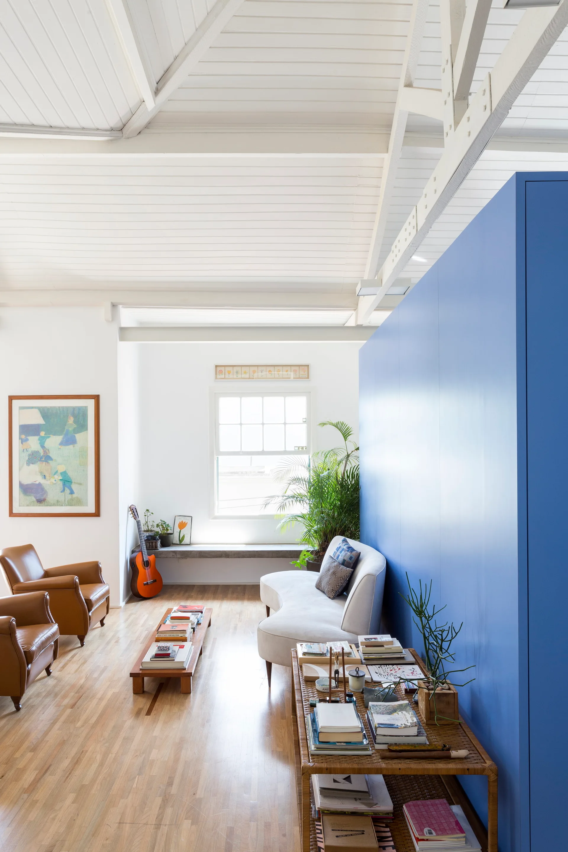 A bright living room features a curved white sofa set against a tall blue partition wall, with leather armchairs and a beamed ceiling.