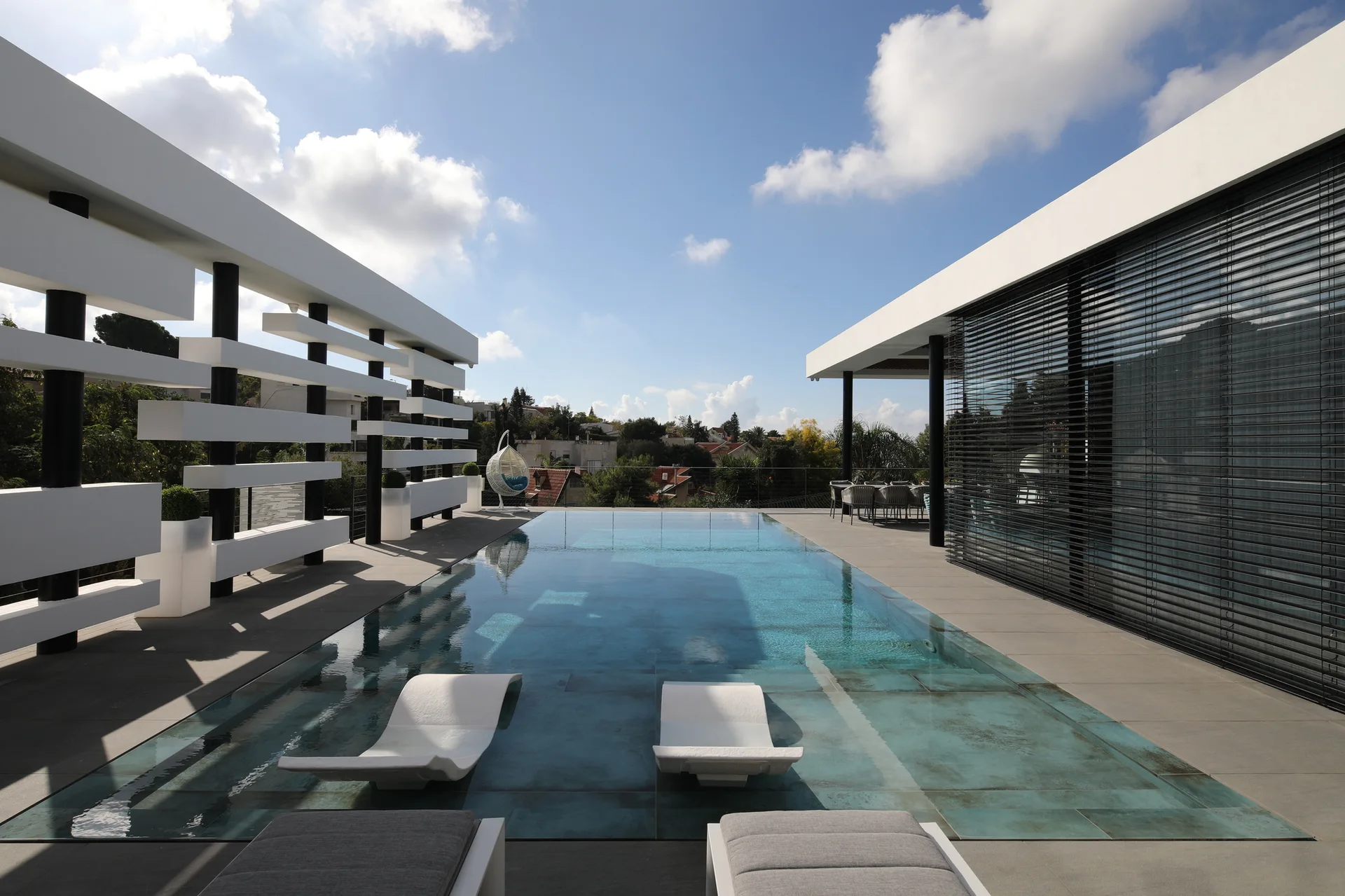 A modern rooftop terrace featuring an infinity pool with submerged white lounge chairs, grey stone decking, and a white slatted architectural screen.