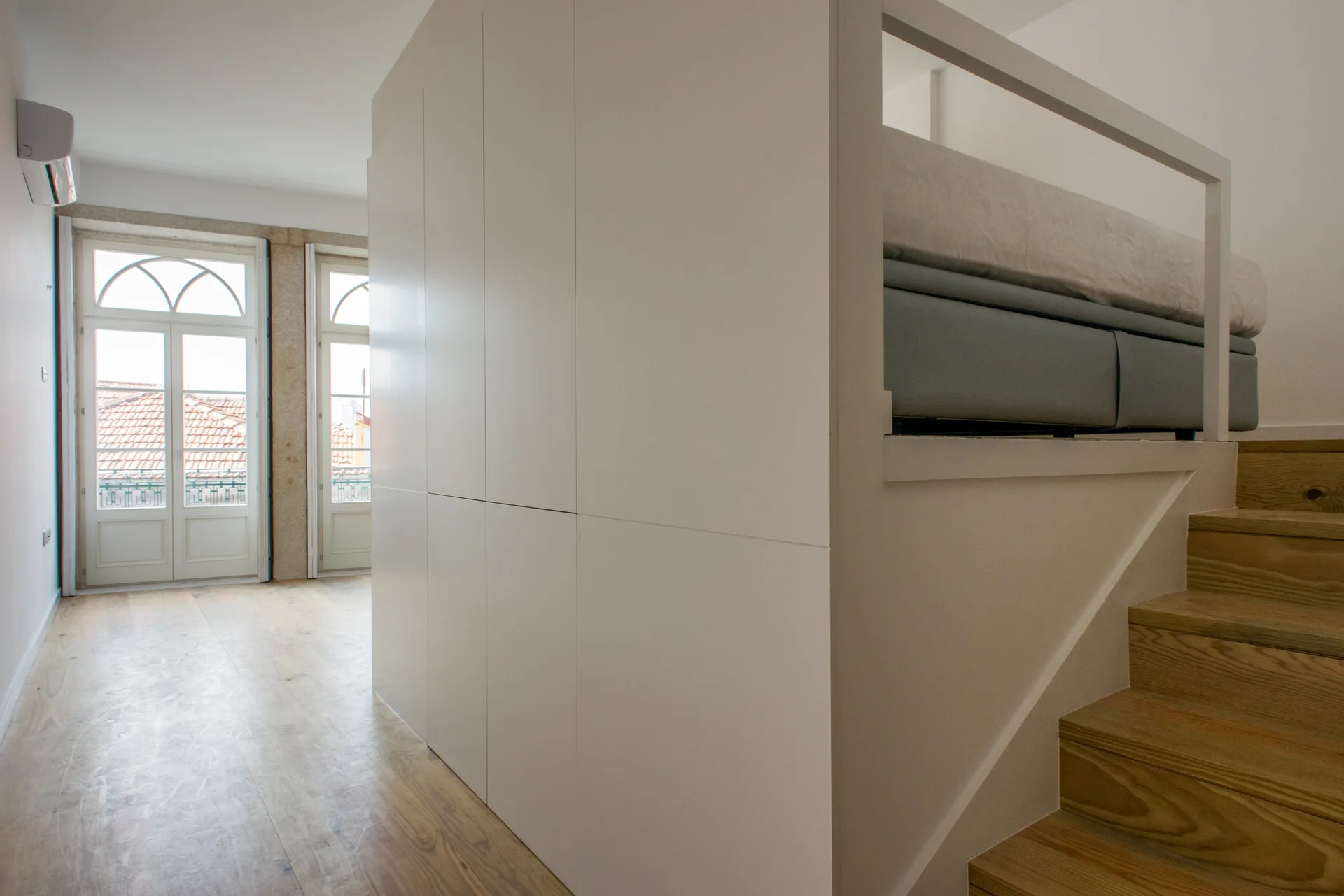 A minimalist loft bedroom featuring a white built-in storage unit with wooden steps leading to an elevated bed next to tall paneled windows.