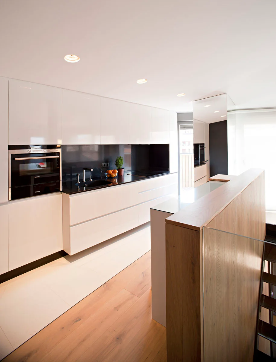 A modern minimalist kitchen features high-gloss white handleless cabinets, a black countertop, and a wood-toned island next to a staircase.
