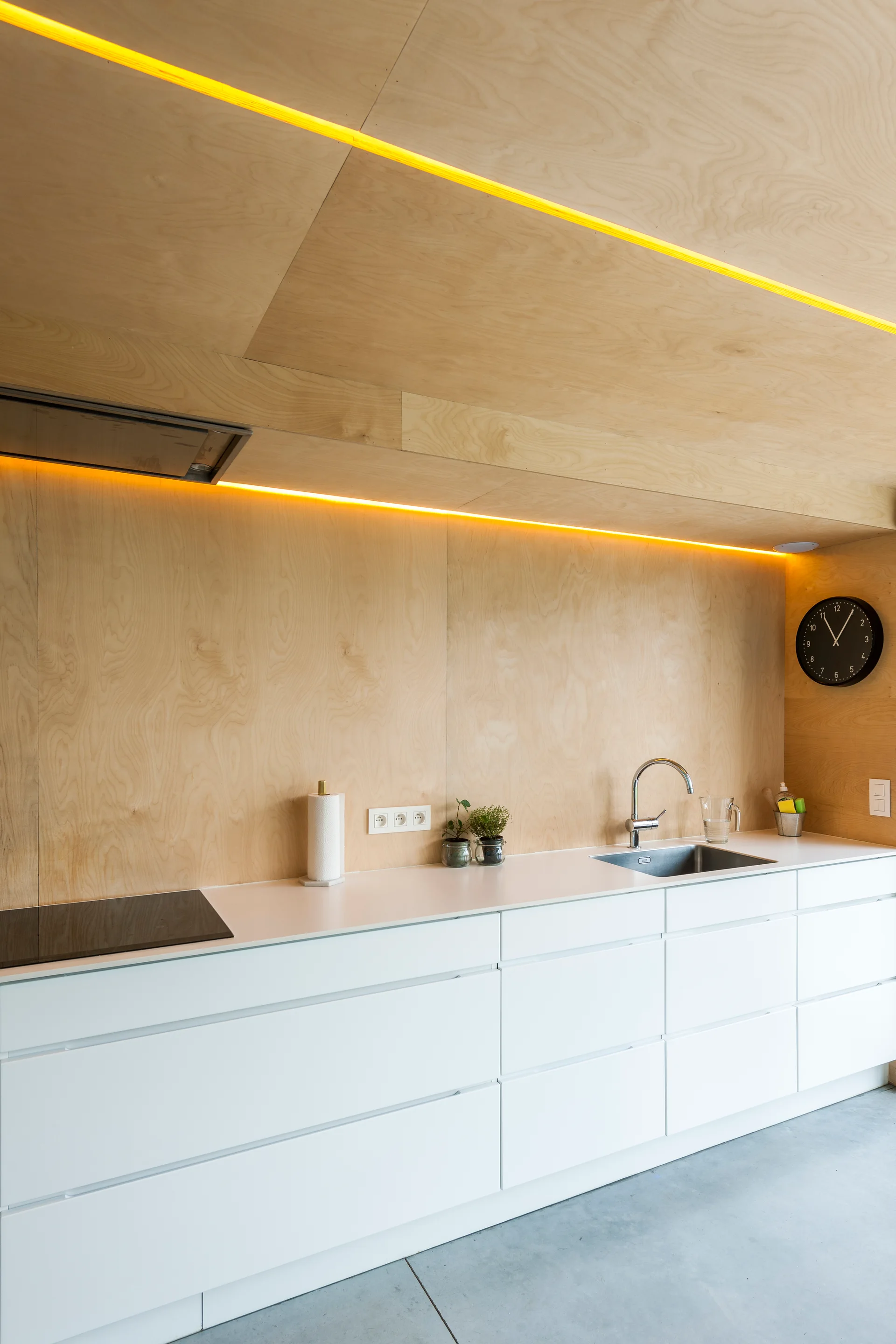 A minimalist modern kitchen featuring light plywood paneling on the wall and ceiling, white handleless base cabinets, and a grey concrete floor.