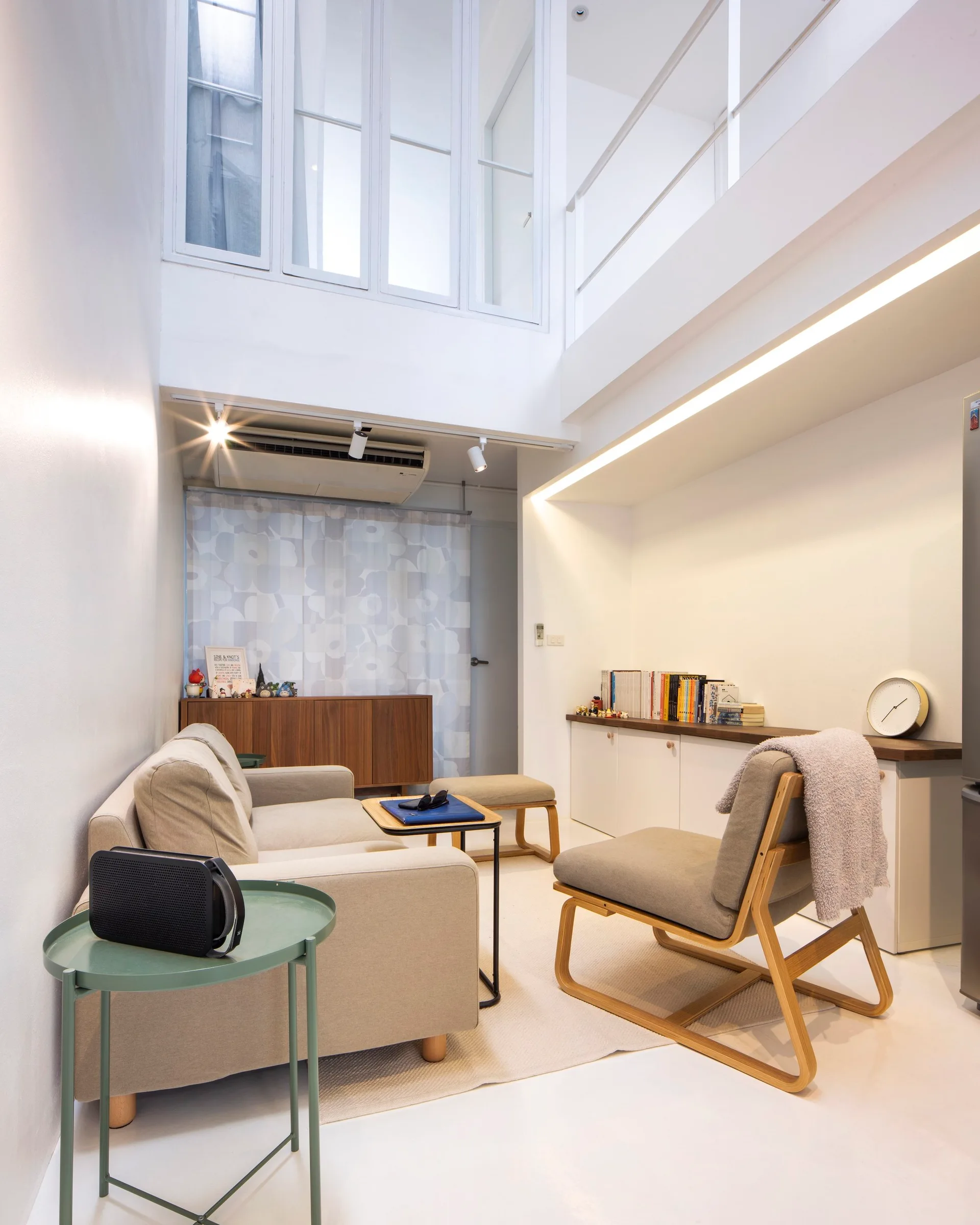 A bright loft living room features a beige sofa, a wood-frame armchair, white built-in cabinets, and high interior windows under a tall ceiling.